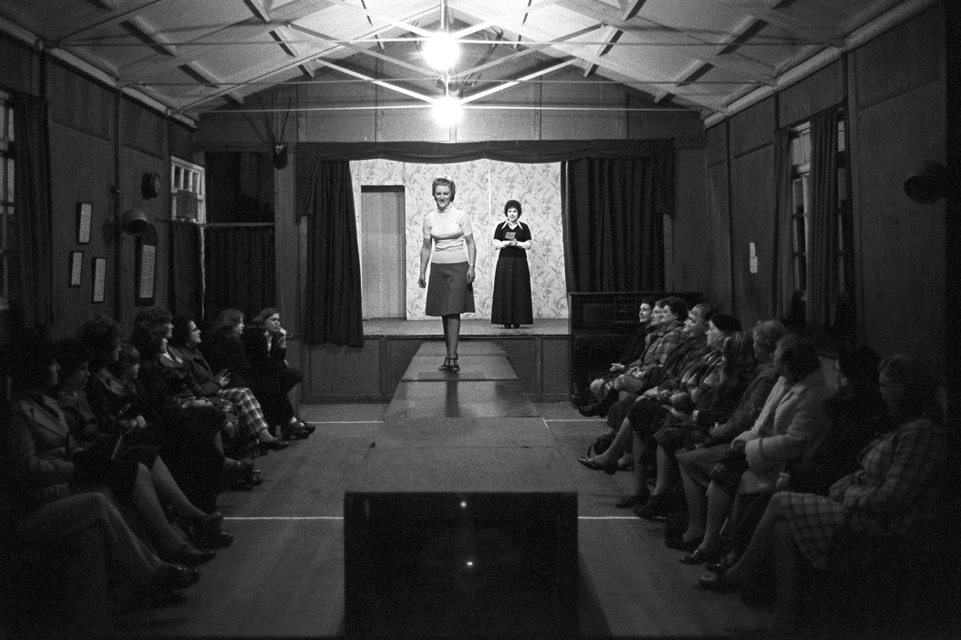Fashion show in village hall (Victory Hall) modelled by women from the village.
[Women watching a fashion show in Beaford Village Hall.  One woman is standing on the stage while another is on the catwalk.]