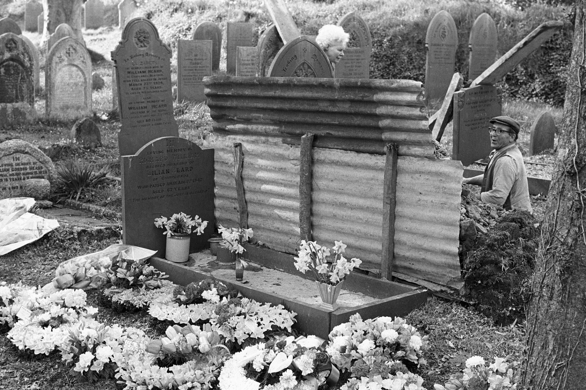 Gravedigger tidying up after burial, flowers in graveyard.
[Gravedigger Reginald Rice tidying Geoff Curtis's grave behind a corrugated iron screen after his funeral in Beaford church.  Another grave and floral tributes are in the foreground.]