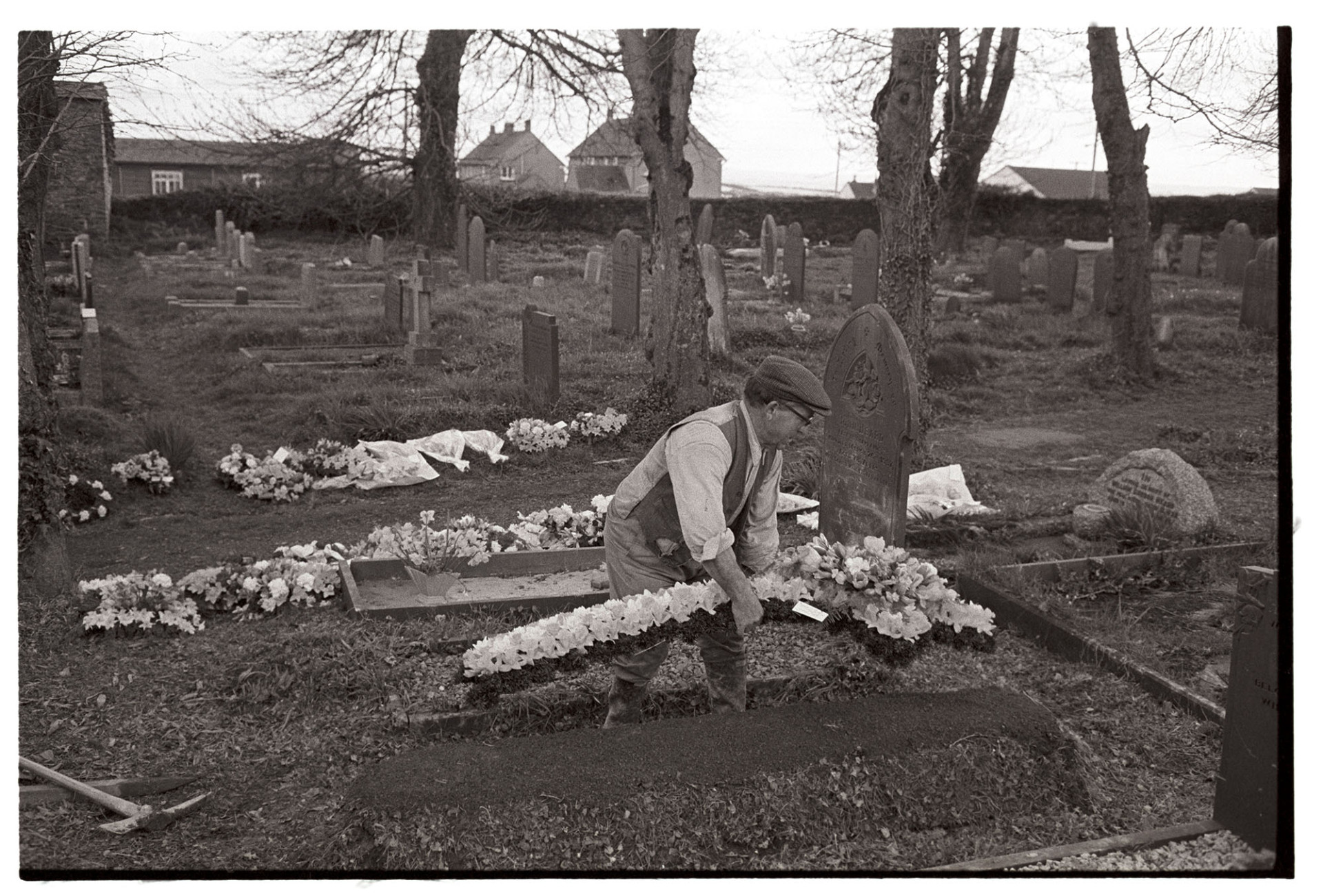 Graveyard with gravedigger tidying up grave after burial. Laying flower cross on grave.
[Gravedigger Reginald Rice laying a large floral cross on a new grave, possibly of Geoff Curtis, which he has just covered in Beaford churchyard.]