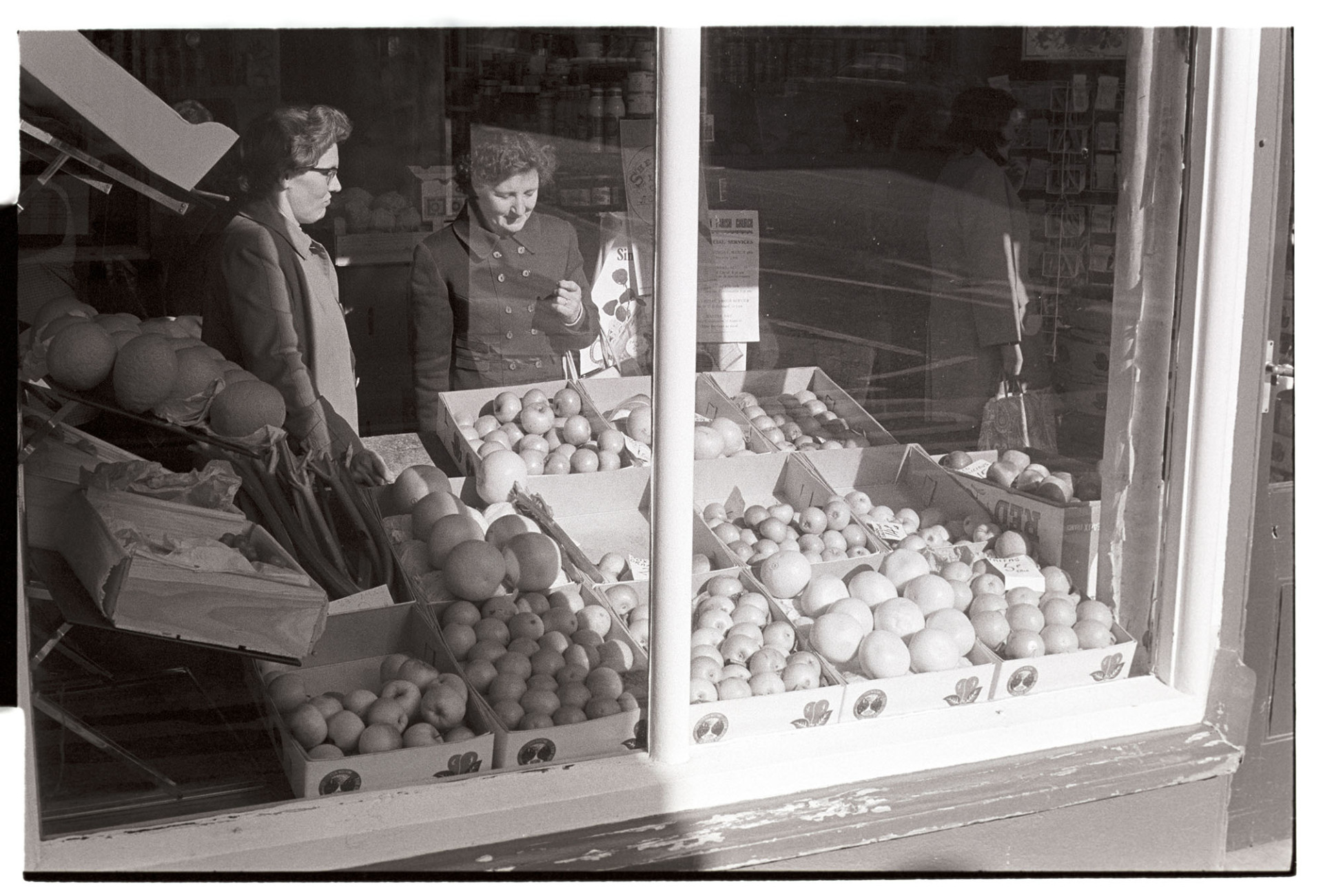 Window of fruit shop with customer.
[Two women, seen through a shop window, inside a greengrocer's shop in Torrington. Fruit, including apples and melons is displayed in the shop window.]