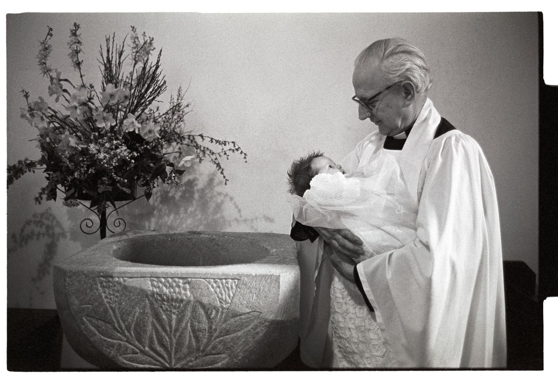Vicar holding baby at font after christening.
[The Reverend Peter Douglas holding Tina Chastey at the font in Inwardleigh church, after her baptism. A flower arrangement is behind the font.]