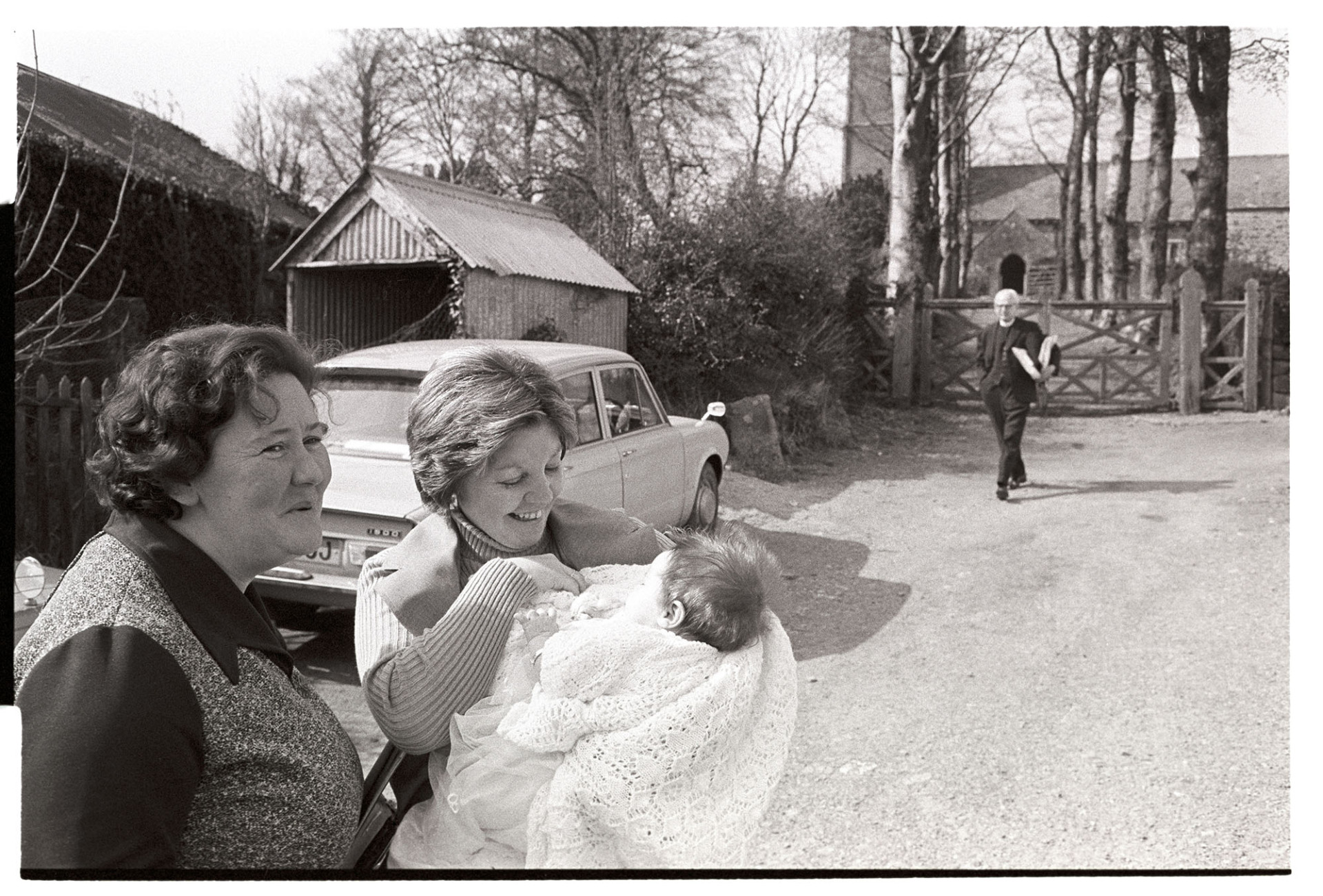 Woman, mother and baby after christening.
[Anne Vallance, stood on the left, with a mother and baby after the baptism of the baby. Inwardleigh church is in the background, with the vicar walking towards them.]