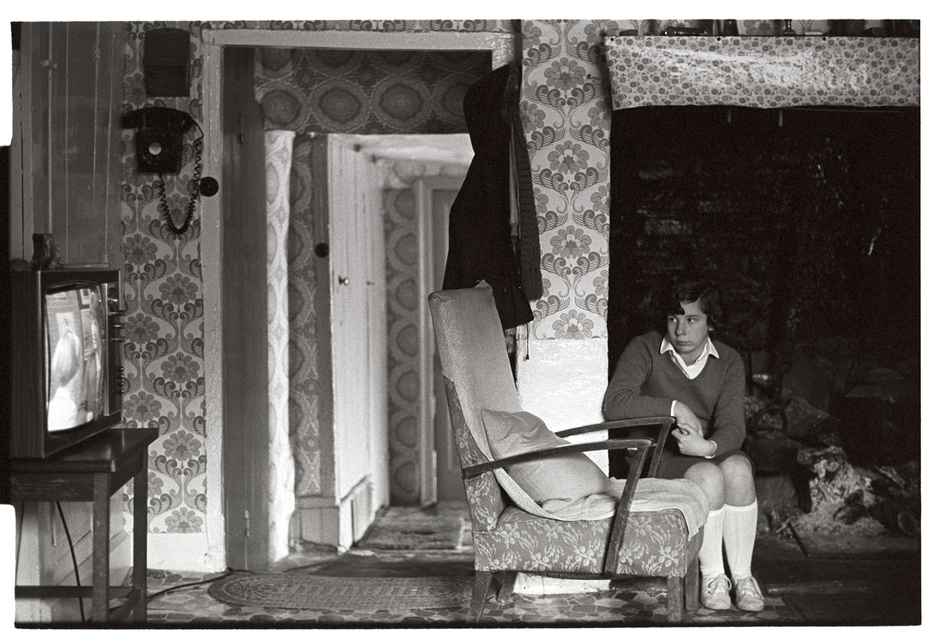 Girl watching TV after school.
[A girl sitting by an open fireplace and leaning on the arm of a chair while watching television after school, in a house at Inwardleigh.]