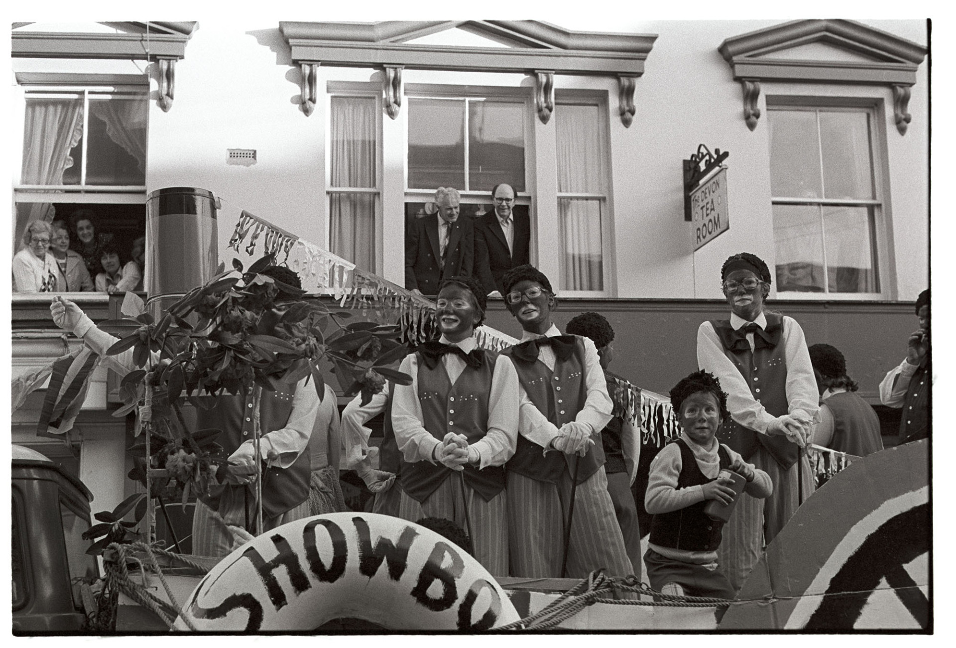 May Fair float. Showboat. Black and white minstrels.
[People dressed as minstrels on a float titled 'Showboat' at Torrington May Fair. Spectators are watching the parade from first floor windows of buildings along the street.]