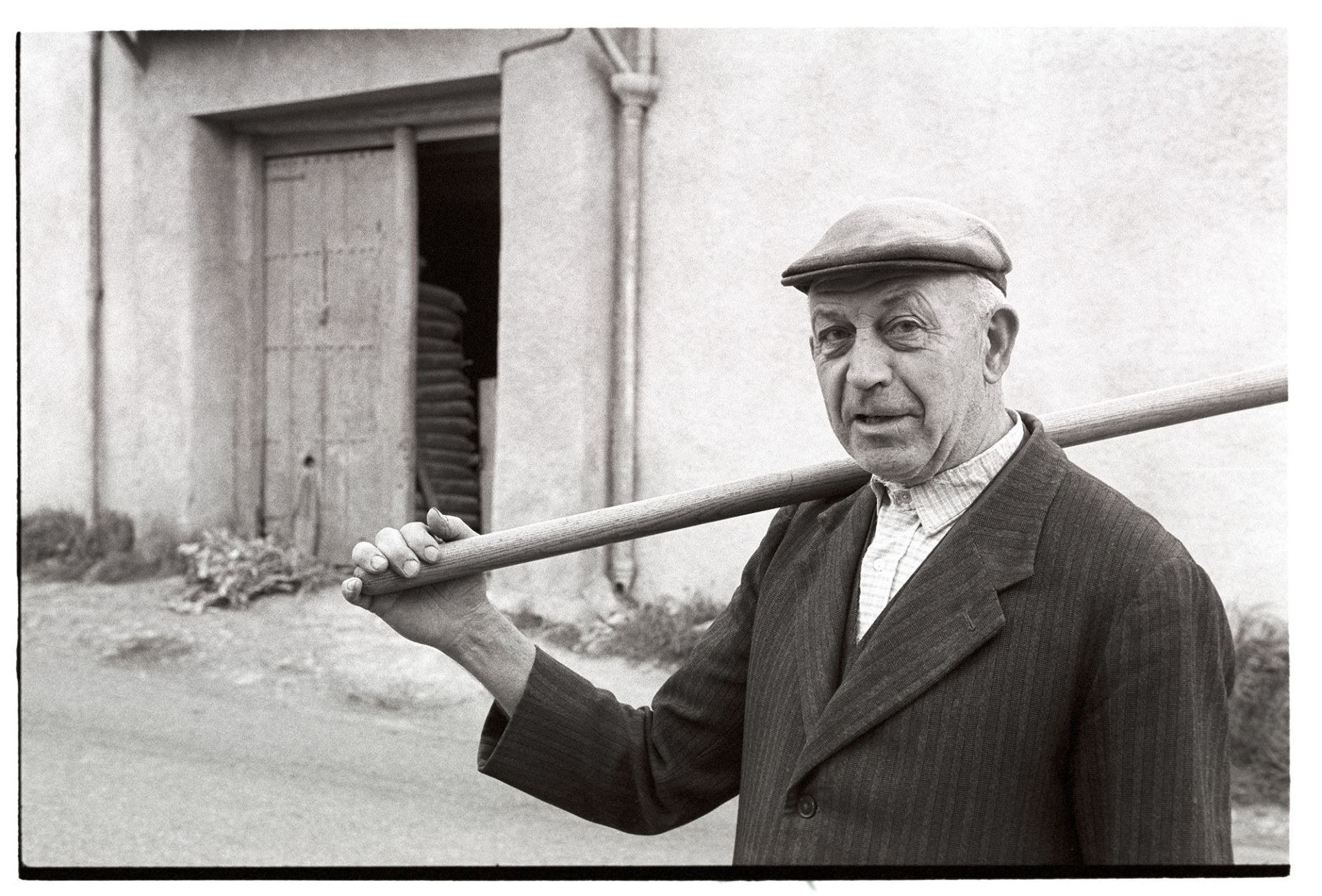 Farmer standing in cap.
[Richard Pearce wearing a cap and carrying a tool on his shoulder and standing in a street in Monkokehampton.]