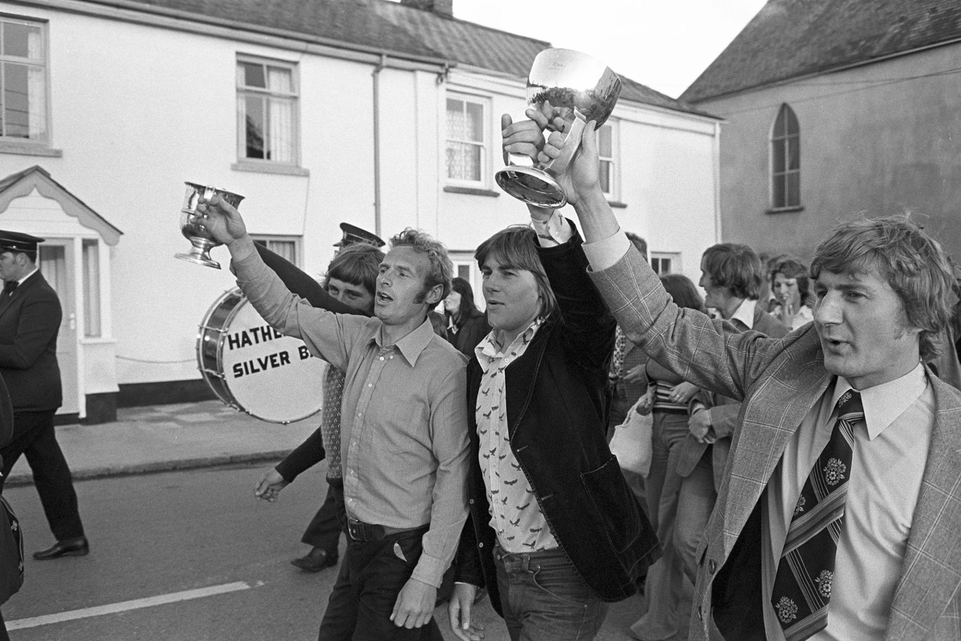 Victory parade with town band to celebrate winning football cup.
[Men parading through a street in Hatherleigh holding aloft silver cups from winning the football cup, with the Hatherleigh Silver Band marching alongside.]