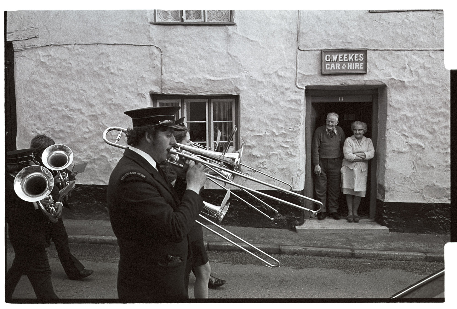 Victory parade with town band to celebrate winning football cup. Taxi Car hire behind.
[Couple standing in a doorway of G Weekes, car hire premises in Hatherleigh,  watching the Hatherleigh Silver Band march past in a parade to celebrate the town winning the football cup.]