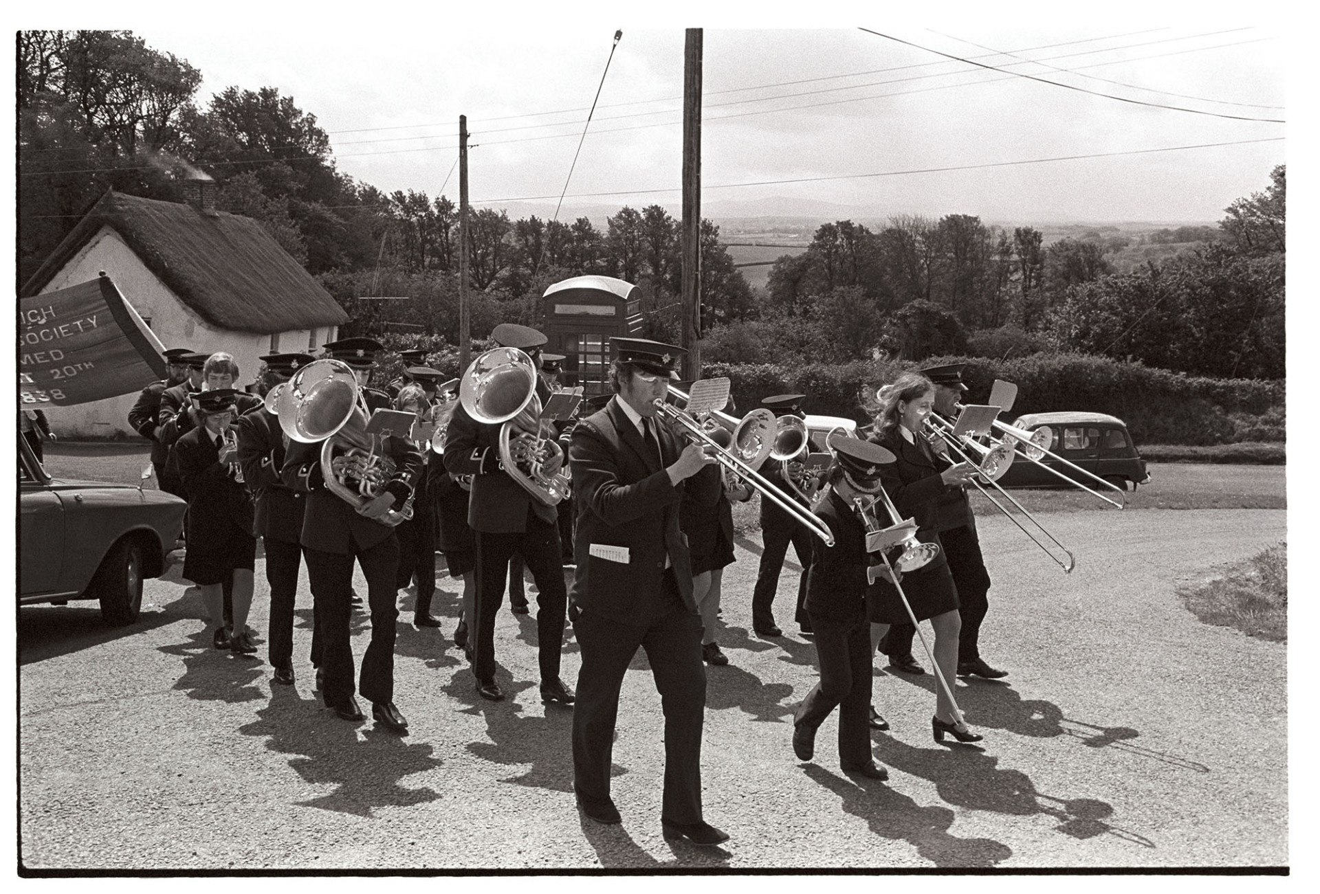 Friendly Society start of parade with Silver Band.
[Hatherleigh Silver Band marching up a road in Iddesleigh for Iddesleigh Club Day. People holding the Iddesleigh Friendly Society banner are following them.]
