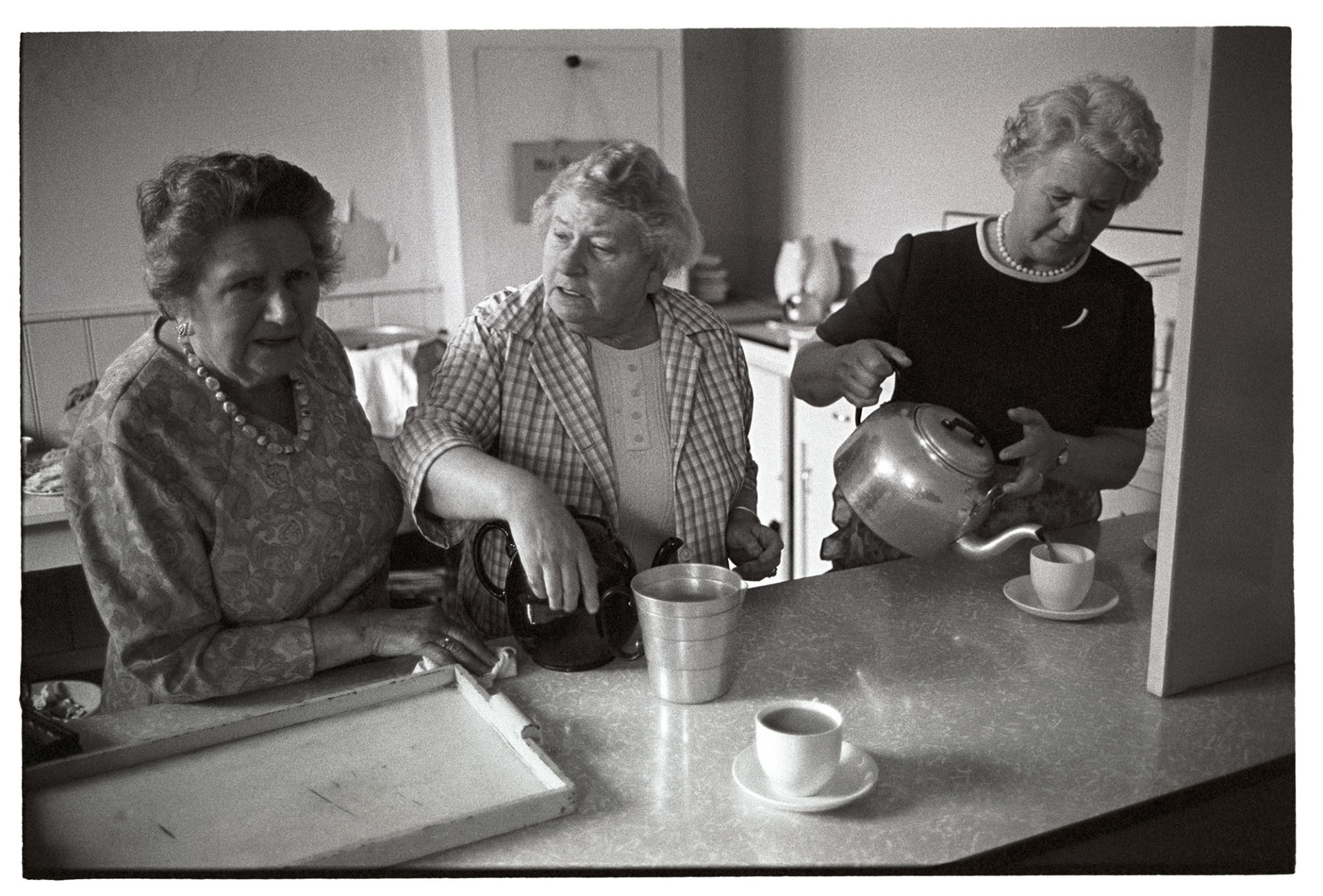Three women serving tea in old school after parade.
[Three women making tea and chatting at a serving hatch, after the Church Sunday School Parade in Hatherleigh.]