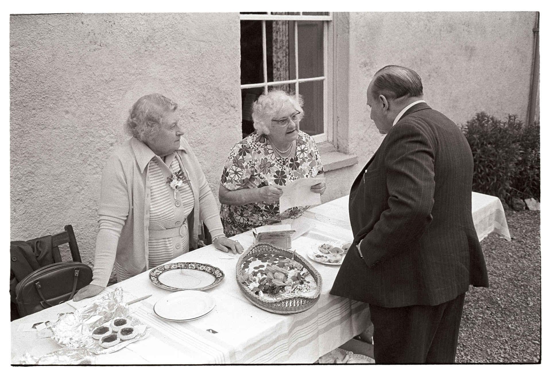 Women and man chatting at cake stall.
[Two women and a man chatting at the cake stall at a garden fete at Reeds, Hatherleigh.]