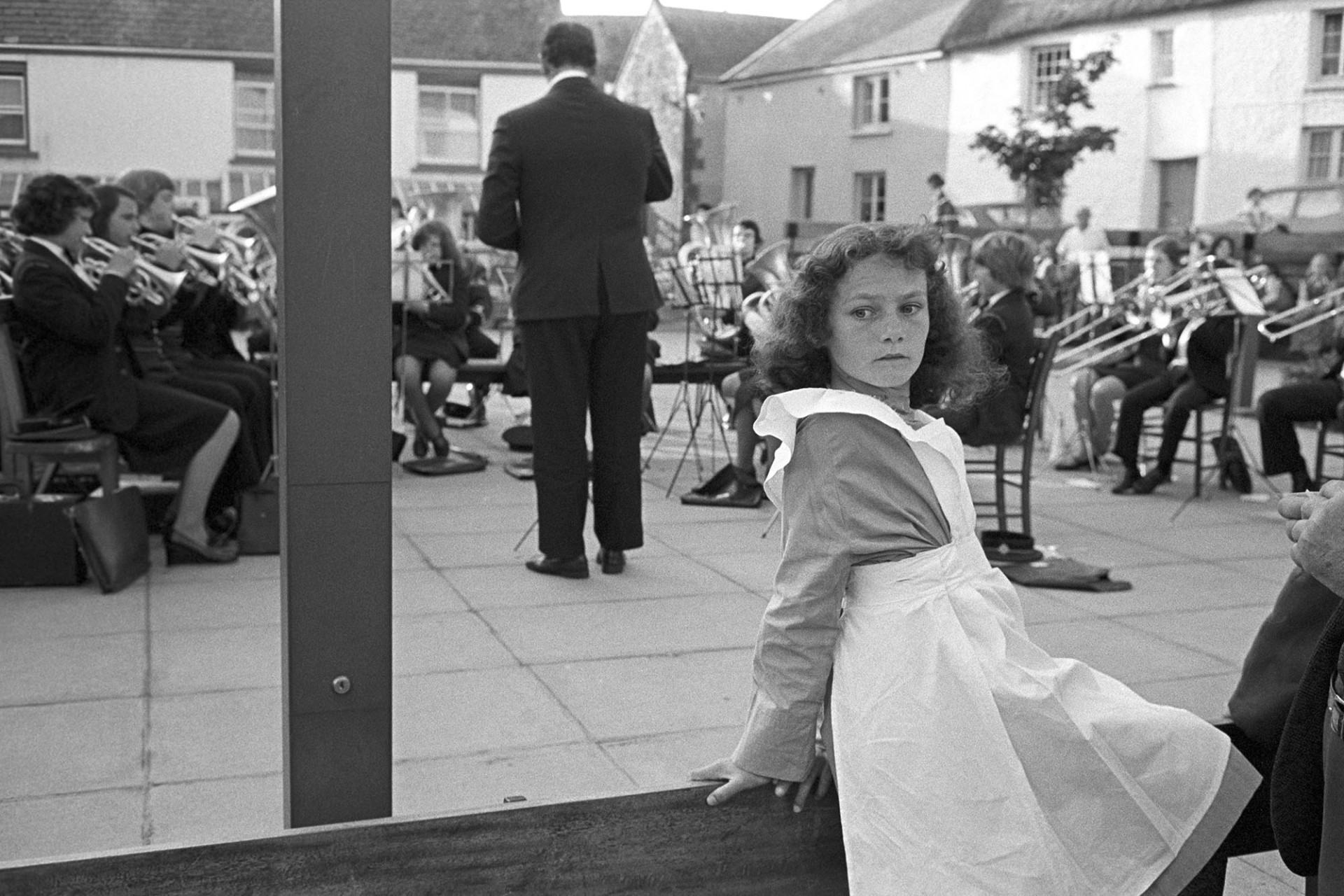 Floral dance. Girl sitting in costume, band playing behind.
[A girl in costume sitting on a wooden fence of railing at Hatherleigh Floral Dance.  The town band is playing in the background in the town square.]
