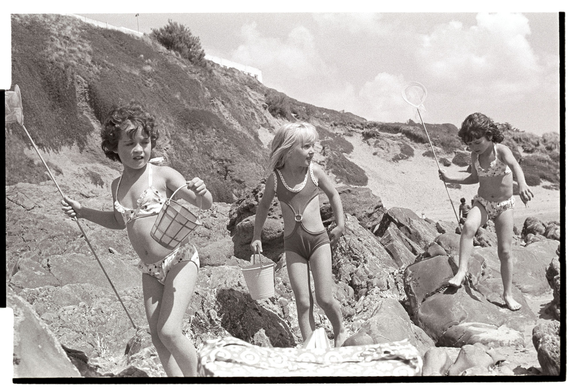 Three children on beach.
[Three young girls with fishing nets and buckets, playing on rocks on the beach, possibly at Instow Sands.]