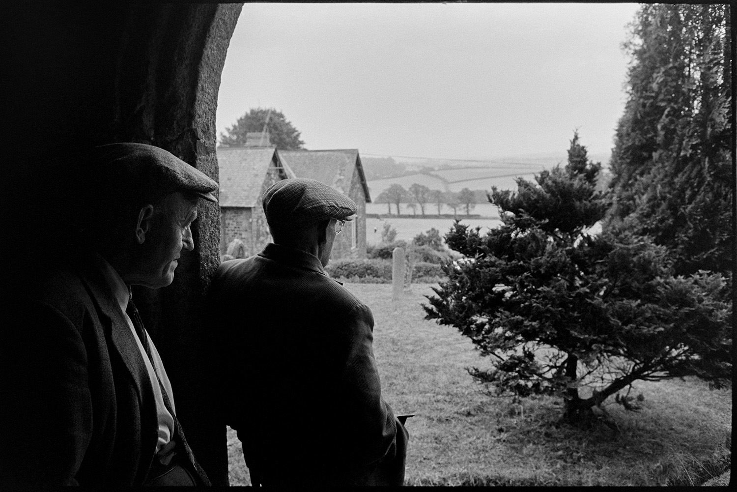 Two men looking from church porch. 
[Two men stood in Iddesleigh Church porch looking out onto the churchyard with gravestones and trees.]
