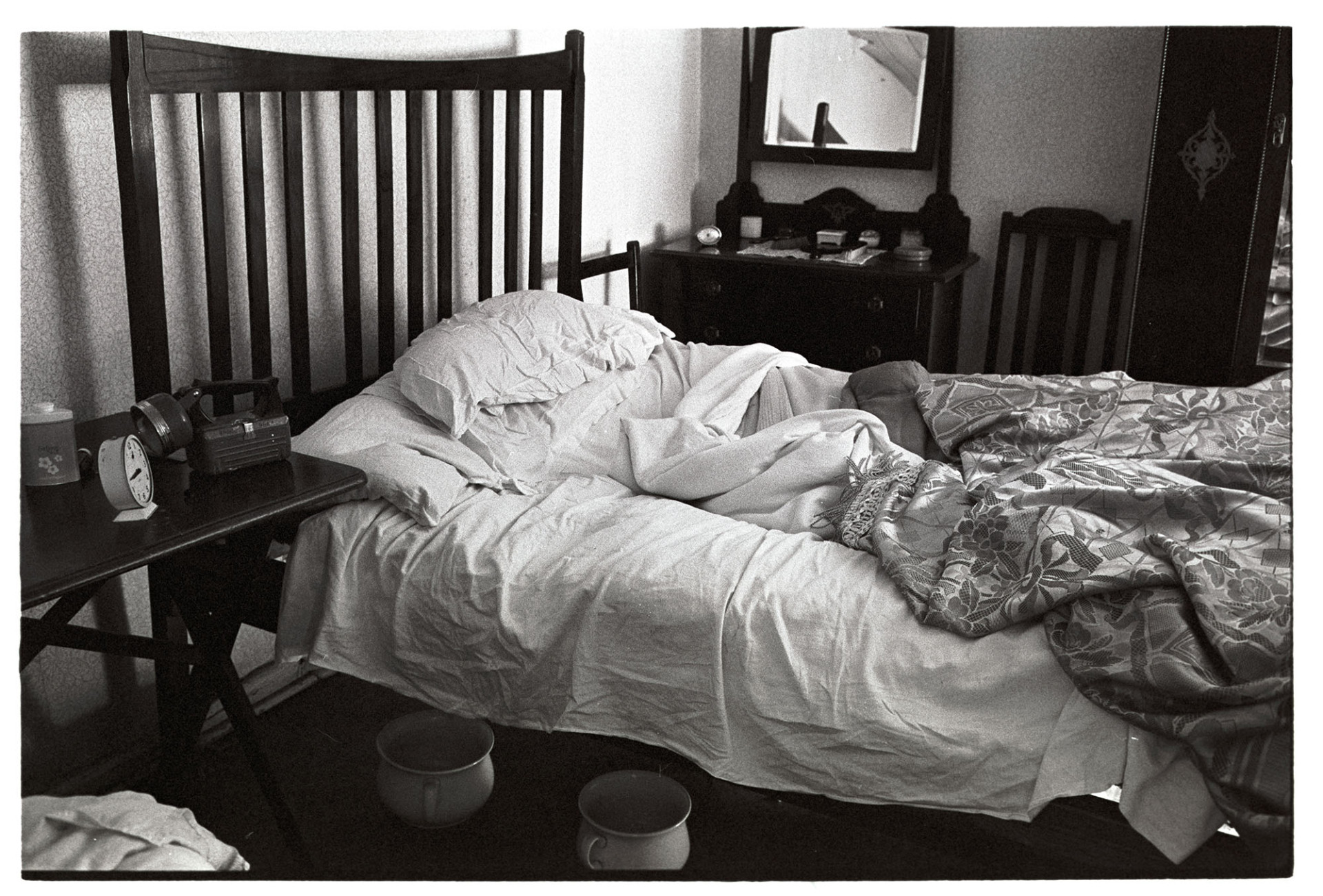 Unmade bed with chamber pots.
[An unmade bed in Archie Parkhouse's bedroom at Millhams, Dolton.  There are two chamber pots beneath the bed, and a dressing table in the background. A torch and alarm clock are on the beside table.]
