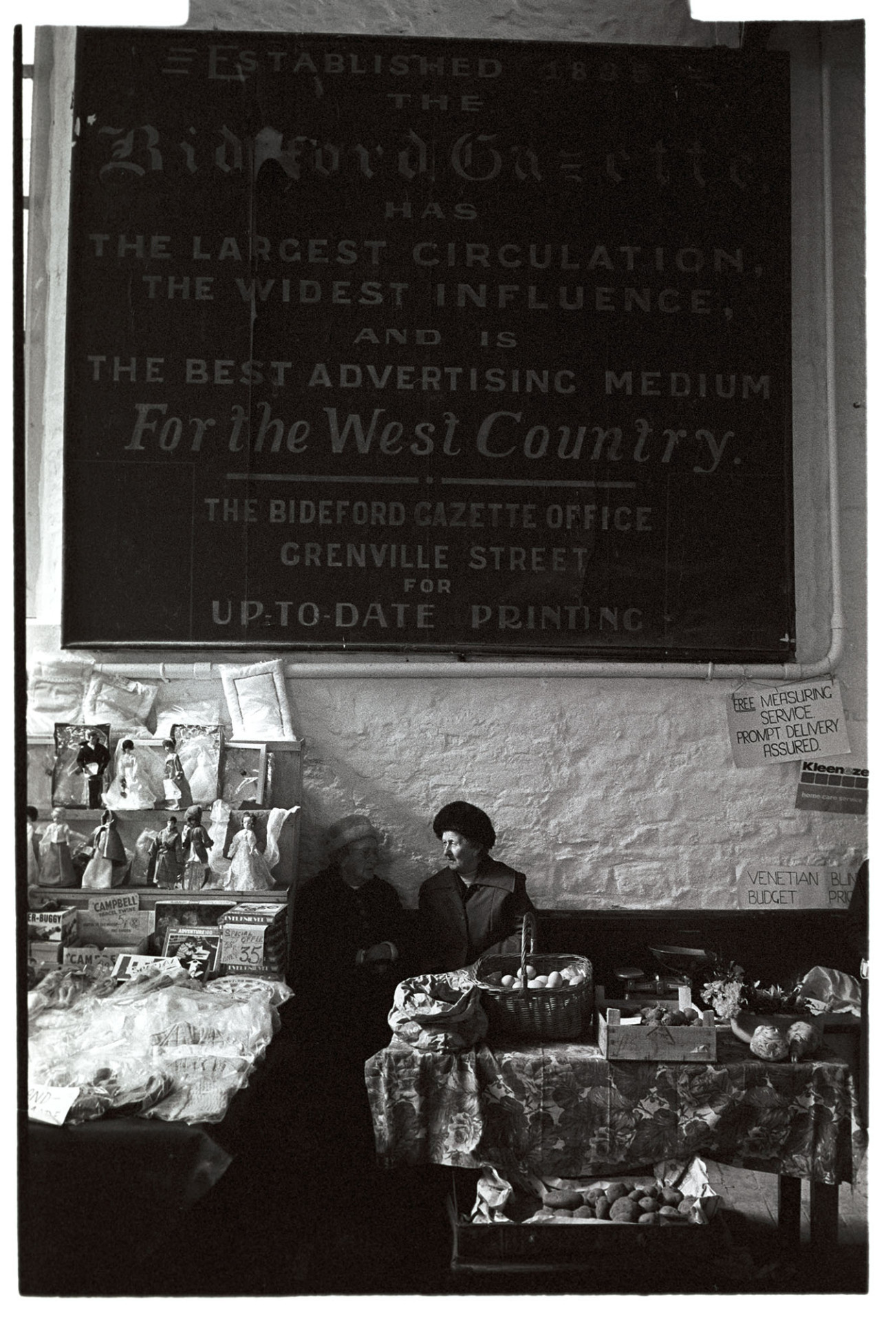 Pannier market. Advert for local paper, dolls boutique.
[A vegetable stallholder and doll stallholder chatting beneath a Bideford Gazette advertisement poster in Bideford Pannier Market.]