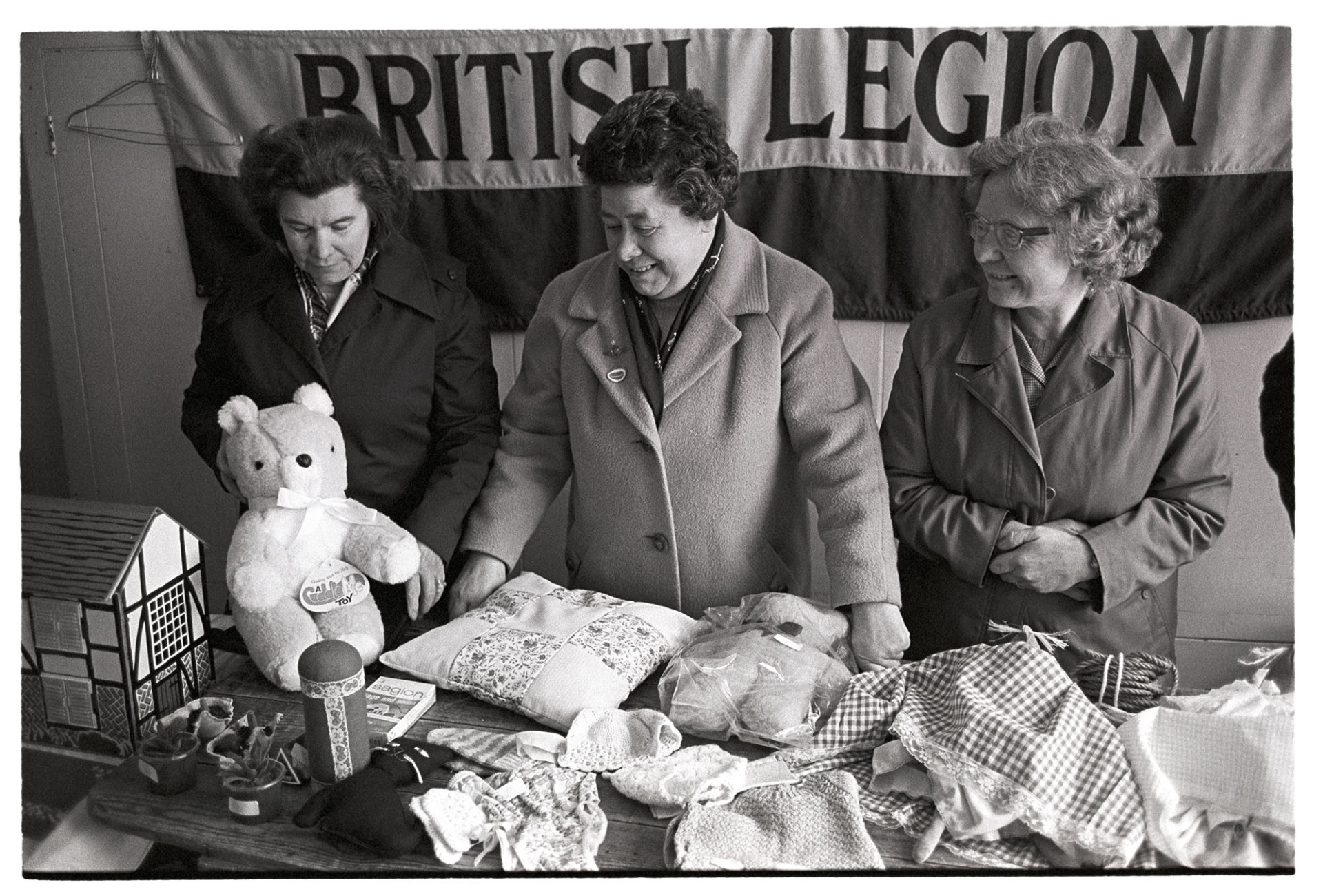 Women at British Legion stall in Pannier Market, toys.
[Three women at the British Legion stall in Barnstaple Pannier Market.  On the stall are soft toys, a dolls' house and soft furnishings.]
