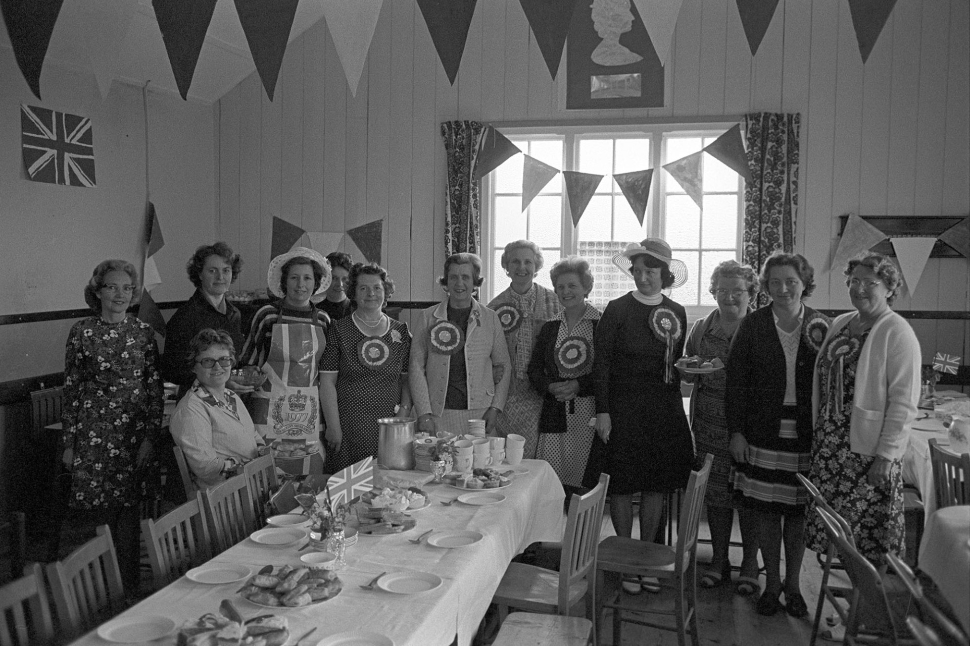 Group of women, in village hall before Jubilee tea which they had prepared.
[Women in High Bickington village hall standing by the tea they have prepared for the Silver Jubilee of Queen Elizabeth II. The tables are laid with cakes and sandwiches and the hall is decorated with bunting and union jack flags. Some of the women are wearing rosettes.]