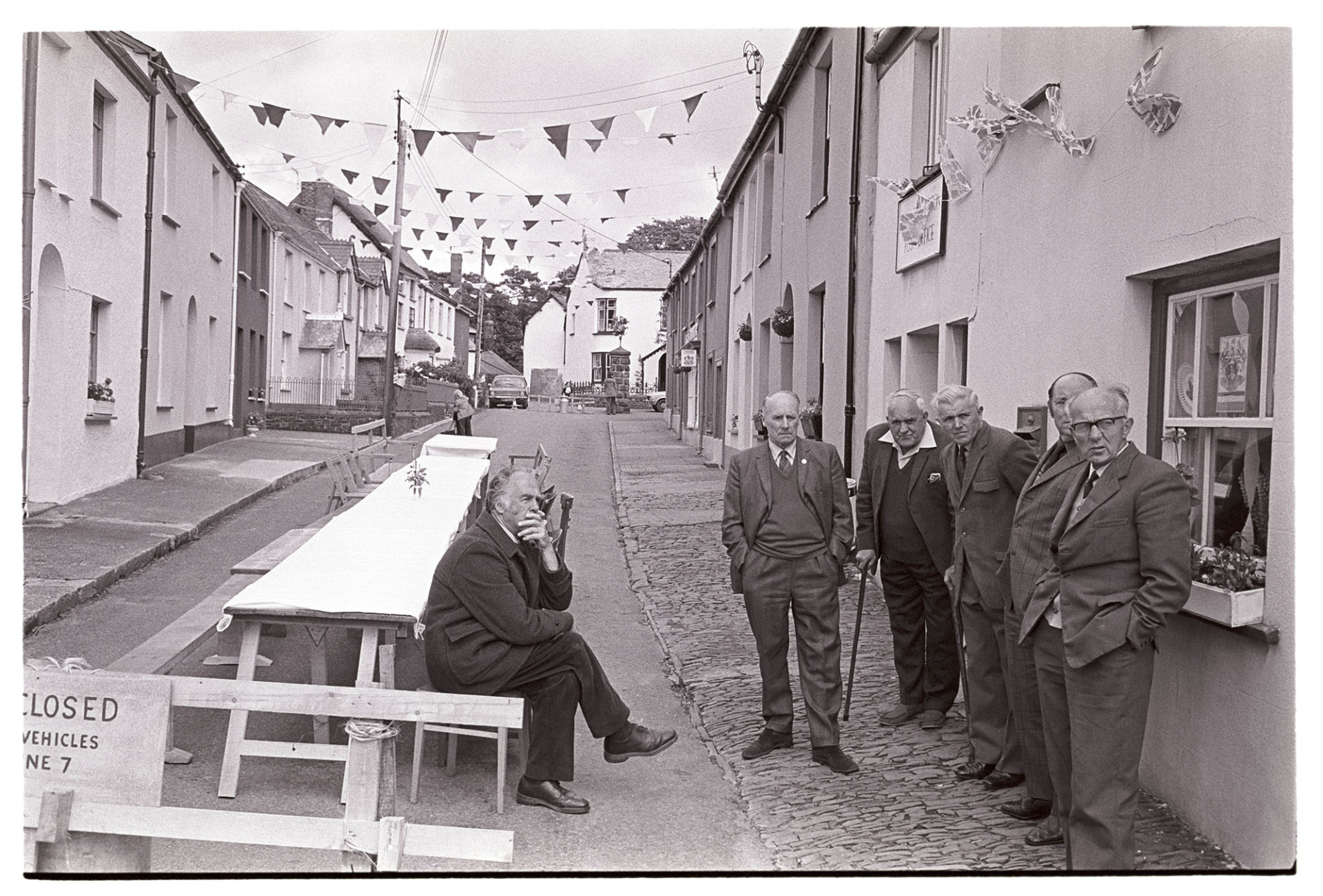 Men waiting for start of Jubilee tea in village street.<br />
[Men waiting in a street decorated with bunting for the Silver Jubilee of Queen Elizabeth II at High Bickington. From left to right the men are Bill Norman (seated), Bill Pidler, Tom Courtney, John Tucker, Bill Underhill and Wilf Marden. They are waiting for the tea and street party to begin. Some of the men have walking sticks.]