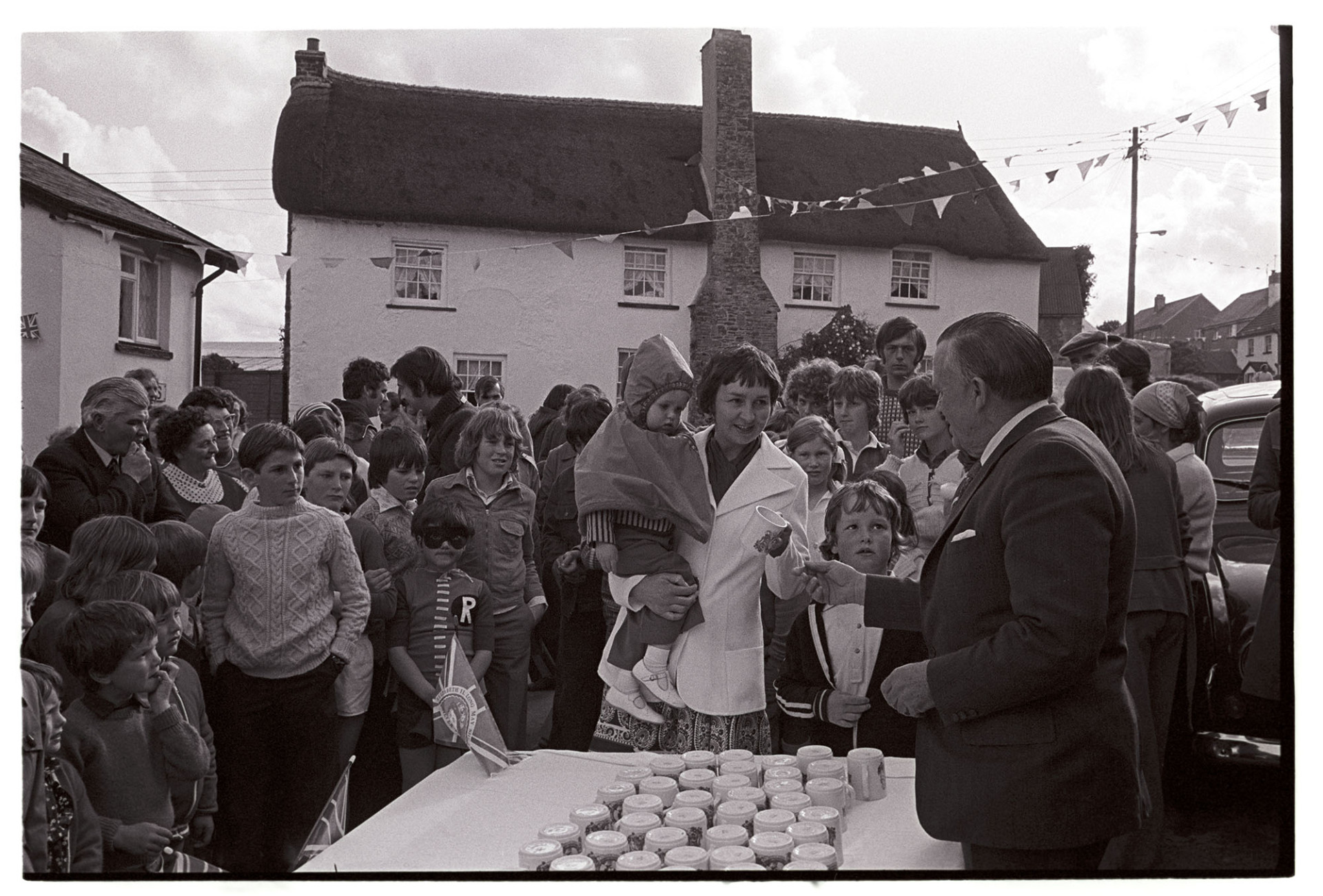 Presentation of Jubilee mugs, crowd in front of mugs set out on table, man presenting them.
[A man presenting Jubilee mugs, to celebrate the Silver Jubilee of Queen Elizabeth II, to a crowd of children at Atherington. The street is decorated with bunting and a thatched cottage can be seen in the background.]