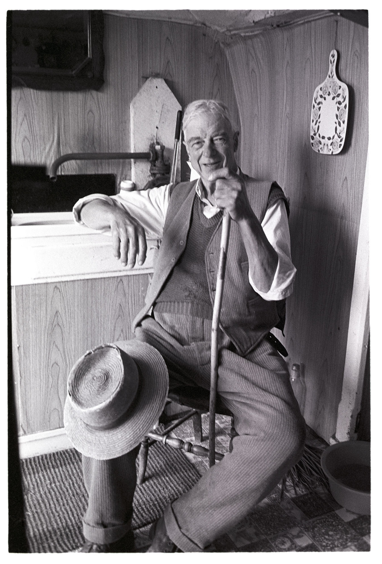Man seated with hat on knee.
[Mr Crocker sitting in the kitchen at Deckport Farm, Hatherleigh holding a walking stick and with a hat on his knee.]