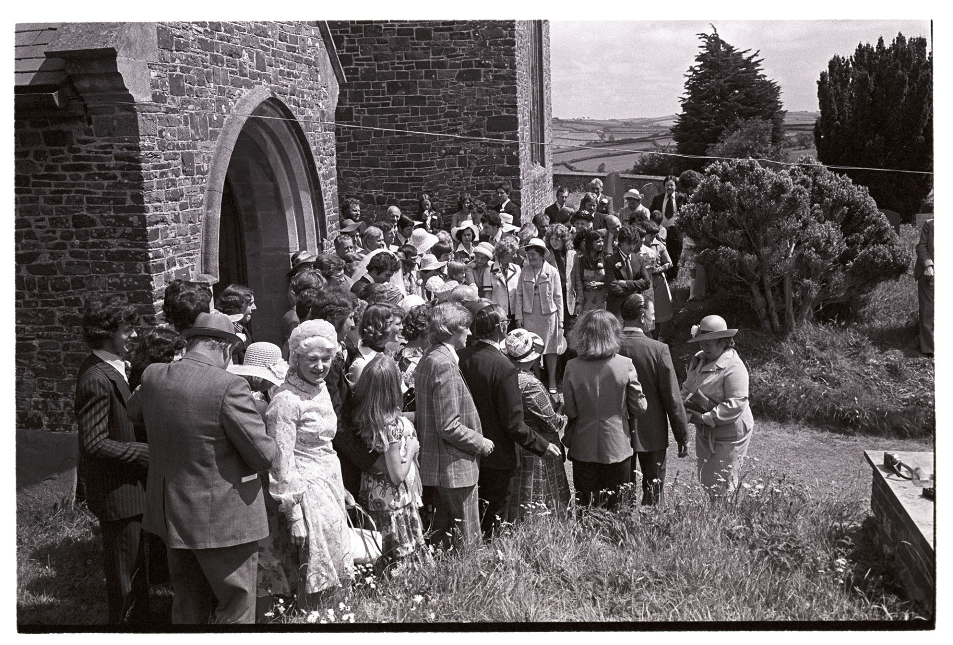 Crowd of guests outside church after wedding. 
[A large group of wedding guests outside the main entrance to the church in Atherington watching the bridge and groom leaving the church.]