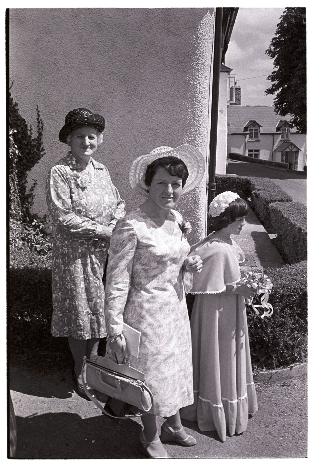 Women guests after wedding in best dresses.
[Three wedding guests stood beside a house in Atherington. Two women are wearing dresses and hats, and a girl, possibly a bridesmaid, is carrying a small bouquet with ribbons.]