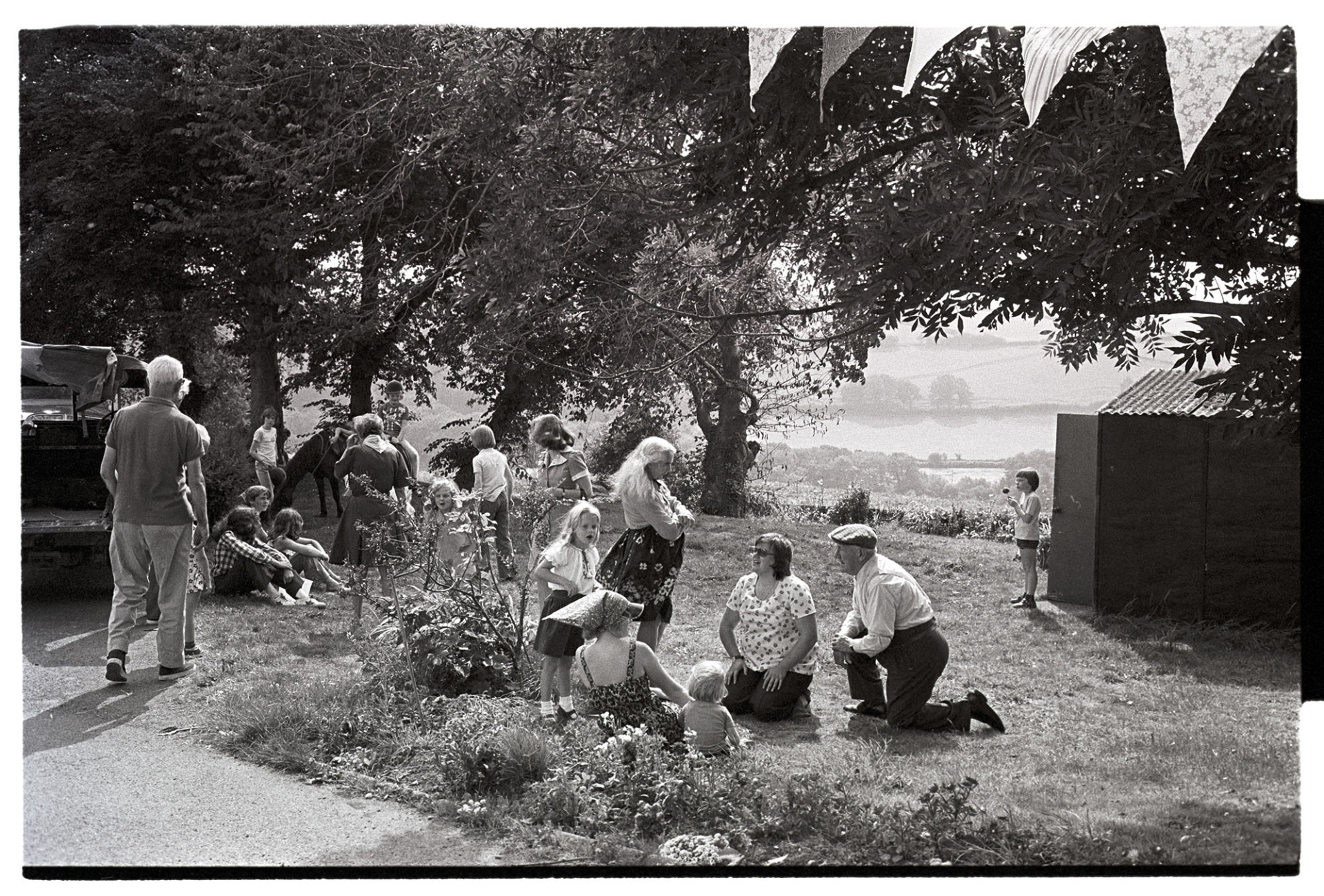 Barbeque fete in grounds of sheltered homes, people sitting out in the sun. 
[Men, women and children sat and stood outside on the grass at a fete at Torridge View, Torrington. There is bunting in the foreground and pony rides in the background.]