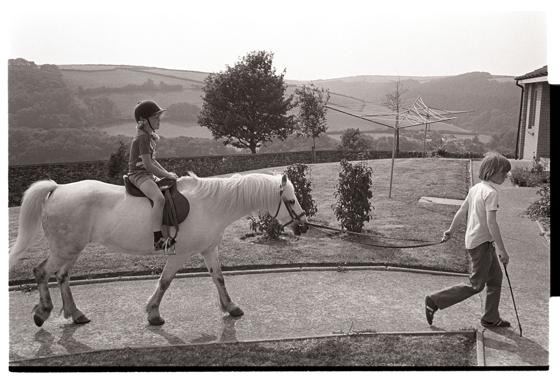 Pony rides for children at barbeque fete in grounds of sheltered homes. 
[A boy in shorts riding on a white pony being lead by another boy at a fete at Torridge View, Torrington. In the background a washing line is visible and a view to fields and hills in the distance.]