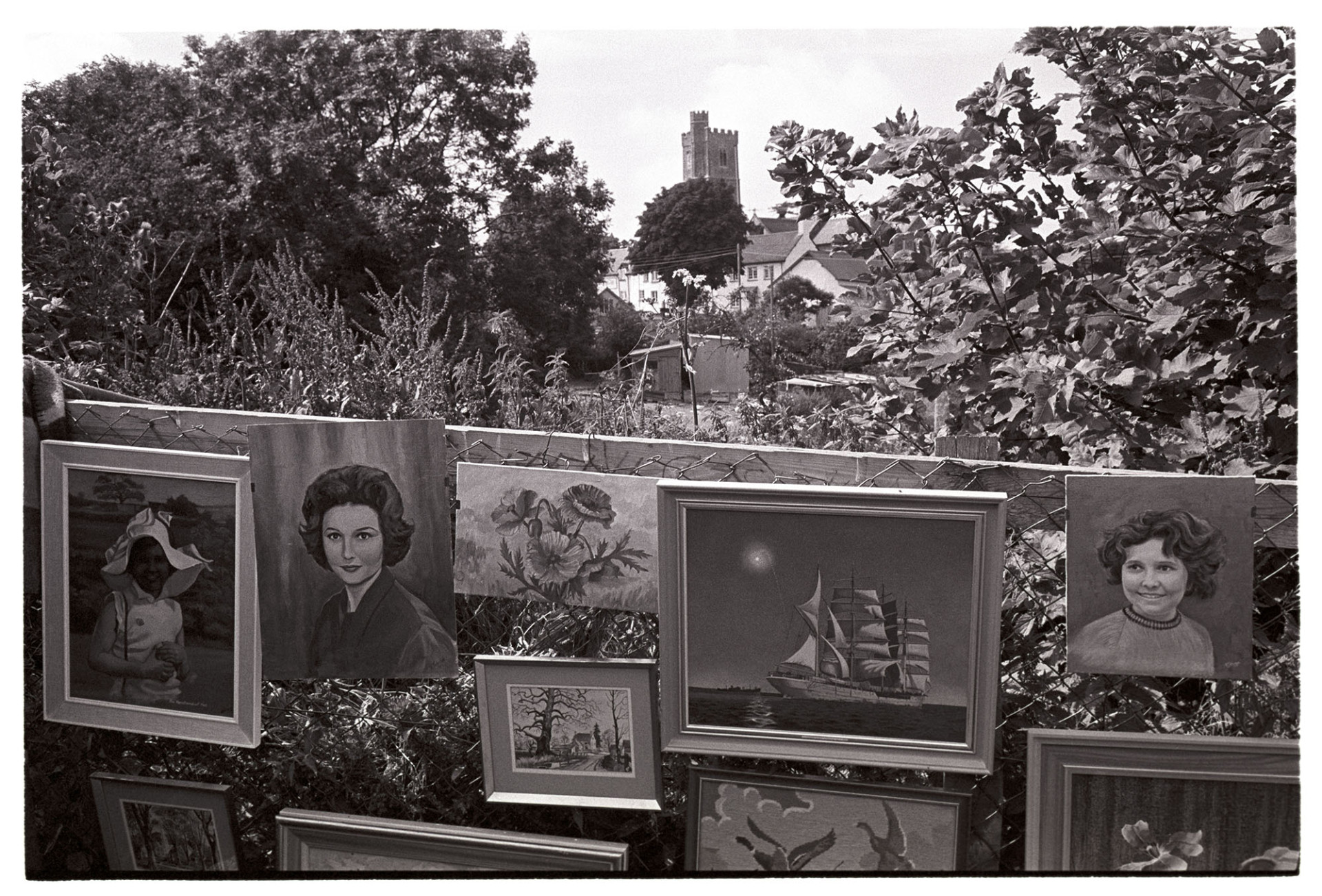 Paintings on show at  fete. 
[Paintings and portraits tied to a fence at the village fete in Atherington. The church and houses are visible in the background.]