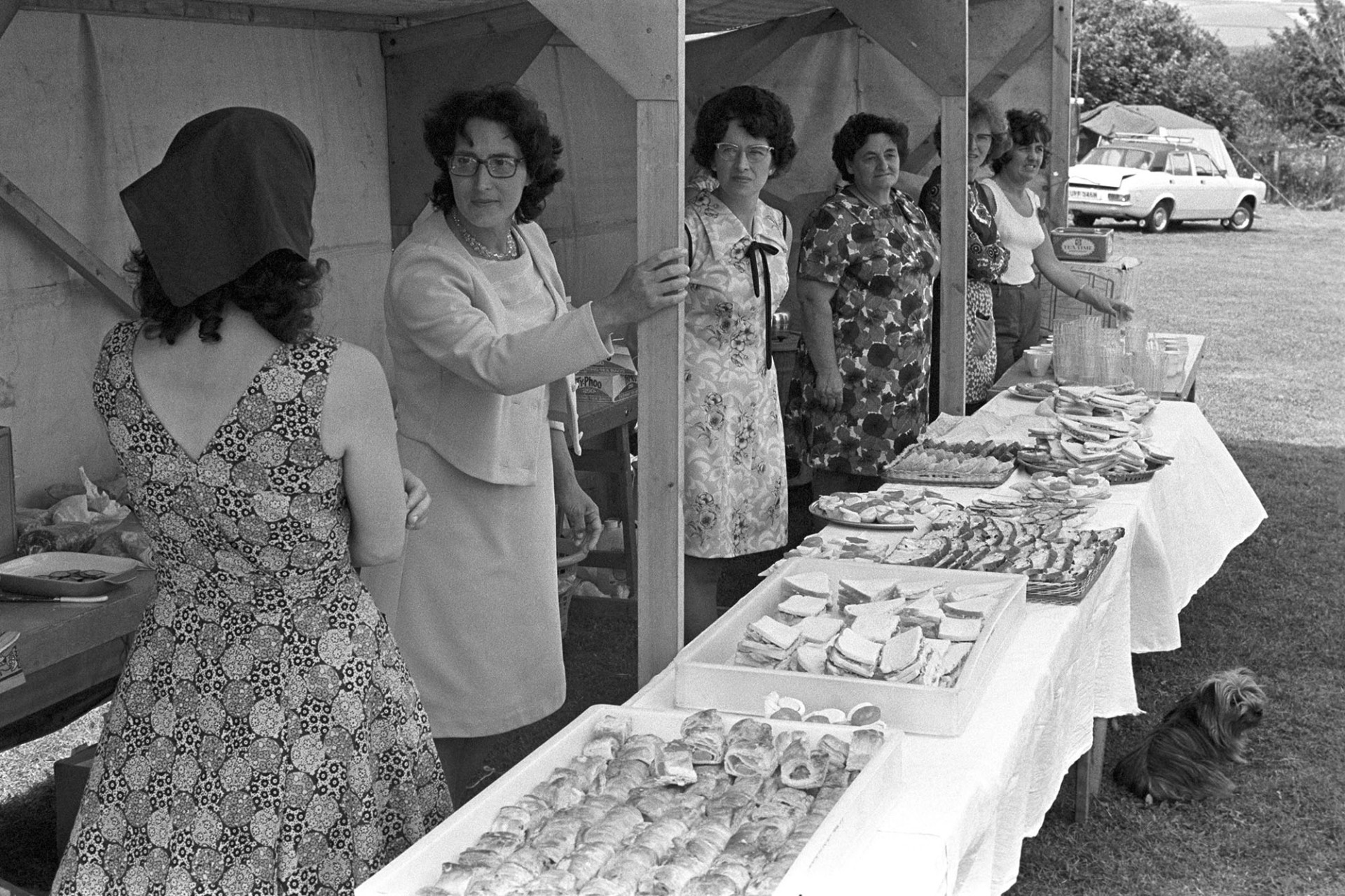 Women at cake stall at fete. 
[Six women at a food and refreshments stall, including sausage rolls and sandwiches, at the village fete in Atherington, with a small dog by the table.]