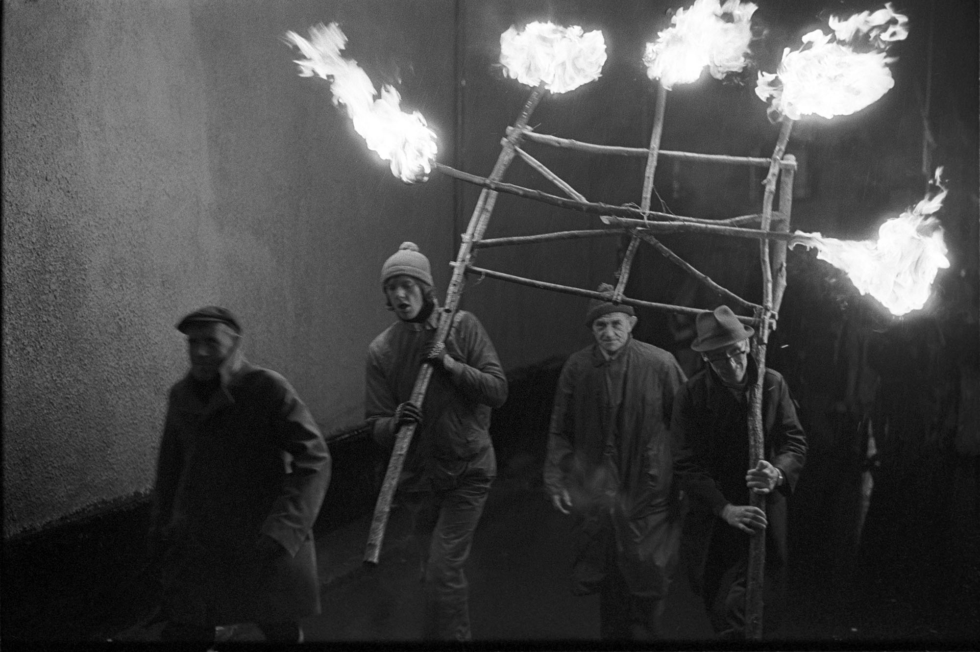 Men carrying frame of flaming torches at start of Carnival.
[Two men carrying a frame of flaming torches at the start of Hatherleigh Carnival.  The frame is made of tree branches. Other people are following in the parade.]