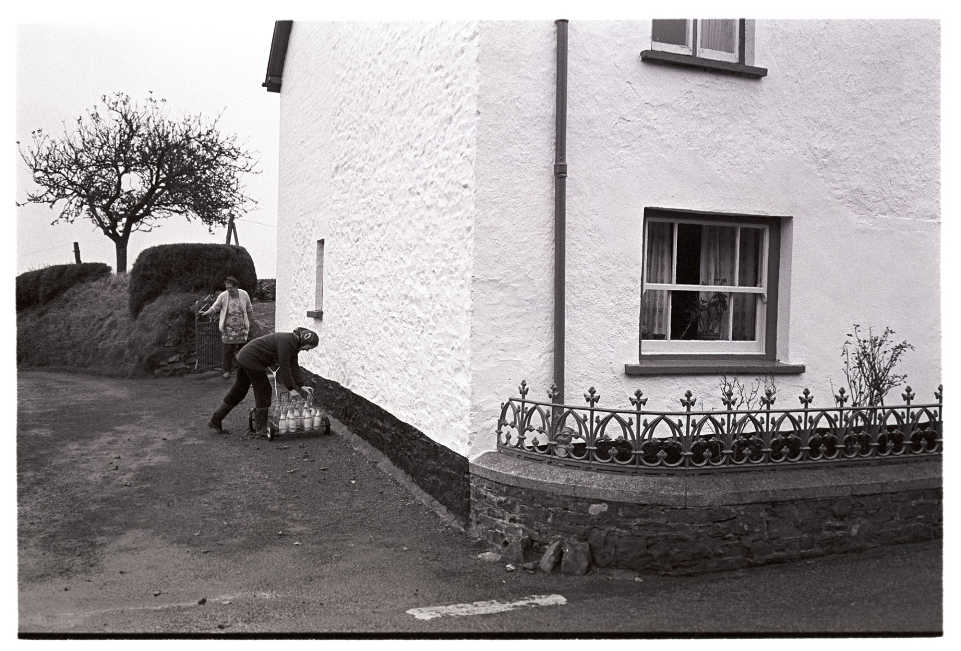Woman delivering milk at house in village with iron railings.<br />
[Jane Woolacott delivering milk from a trolley in Atherington. She is standing beside a house with wrought iron railings.  In the background another woman is emerging from a garden gate.]