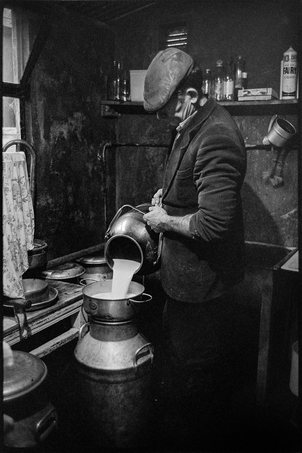 Farmer filling milk churn in dairy. 
[Jim Woolacott filling a milk churn from a smller churn in the dairy at Verdun, Atherington. Other milk churns are visible and a shelf with various bottles can be seen in the background.]