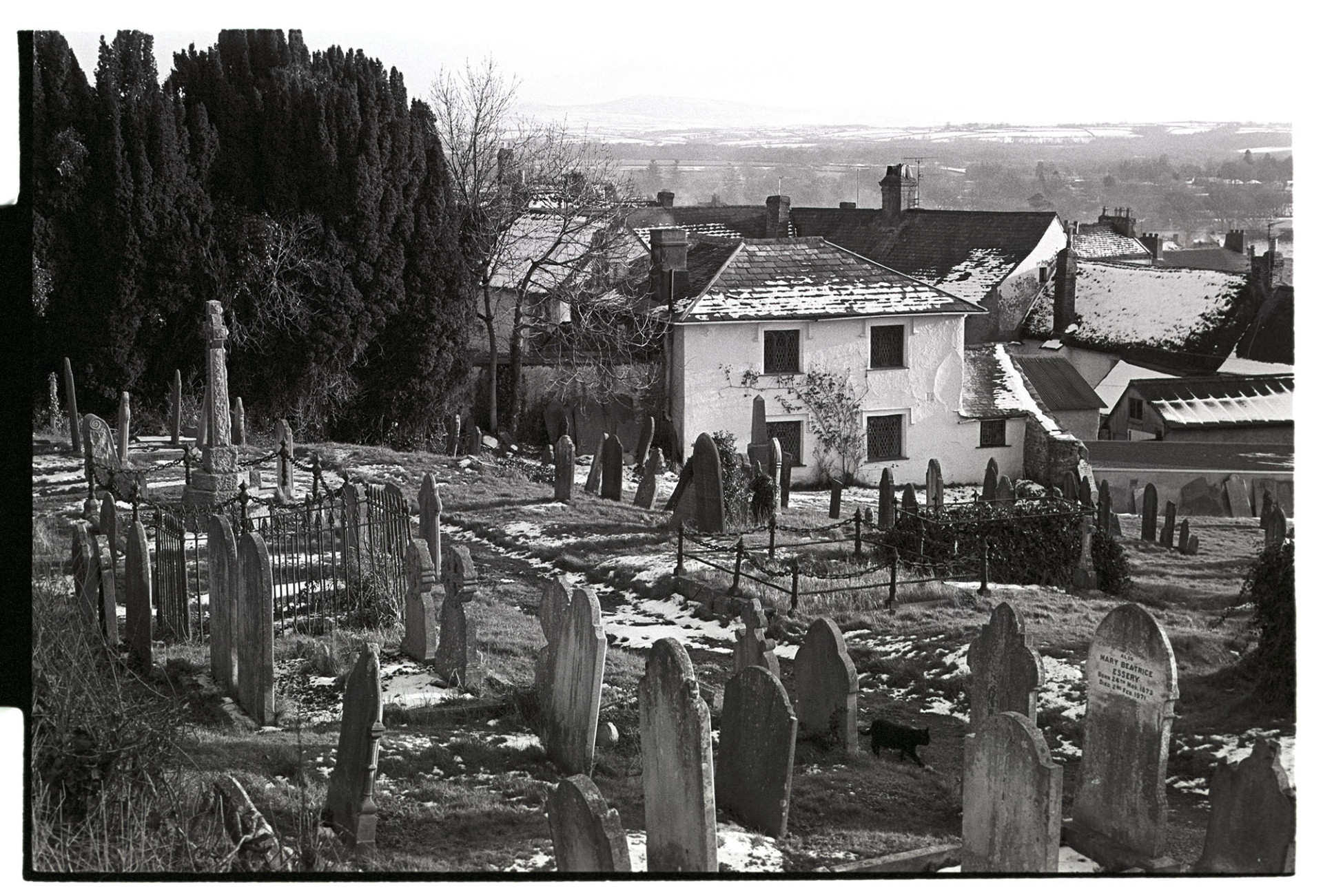 View across graveyard with town under snow, black cat.
[A view of gravestones in Hatherleigh churchyard and snow covered roofs in the town, with Dartmoor in the distance.  A black cat is walking along the churchyard path.]