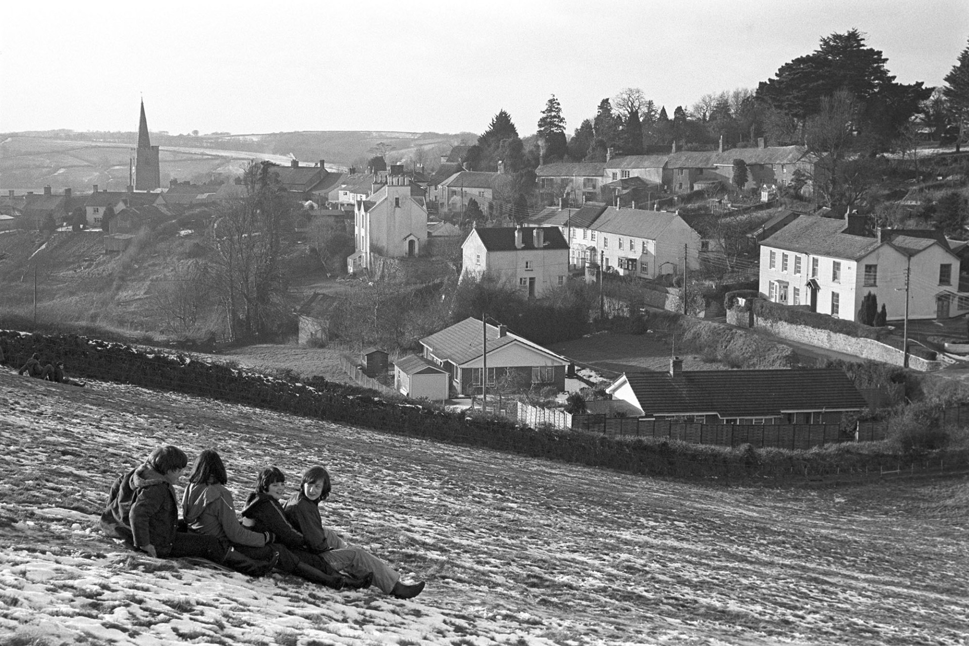 Children tobogganing in front of town, snow.
[Four children tobogganing down a field at Hatherleigh. The town and church tower are visible in the background.]
