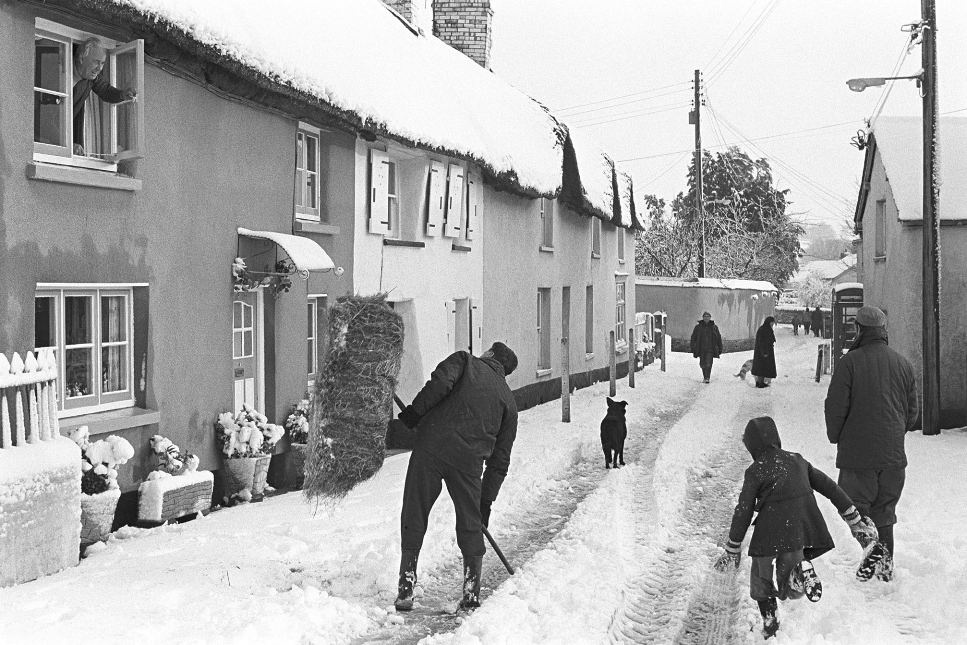 Snow, man taking hay through village to fields, woman with dog.
[A man carrying a bale of hay, possibly Harold Nott, in Fore Street, Dolton, on his way to feed sheep in the snow. He is talking to a man leaning out of a cottage window. A man and child are walking past in the snowy street. Further down the street are a dog and several people walking through the snow.]
