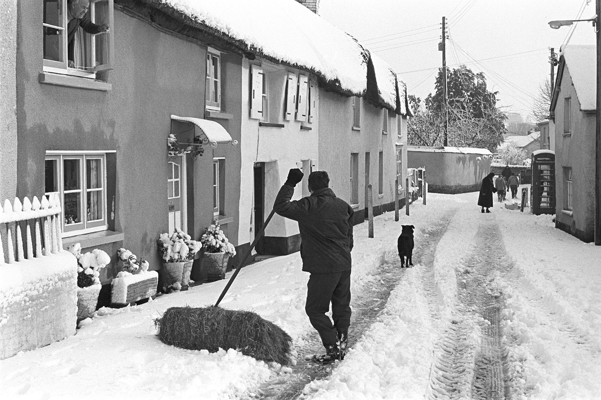 Snow, man chatting in street on the way to sheep with hay.
[A man with a bale of hay, possibly Harold Nott, standing in front of a thatched cottage in Fore Street, Dolton, on his way to feed sheep in the snow. He is talking to a man leaning out of a cottage window. Further down the street are a group of people and dog walking through the snow, past a telephone box.]