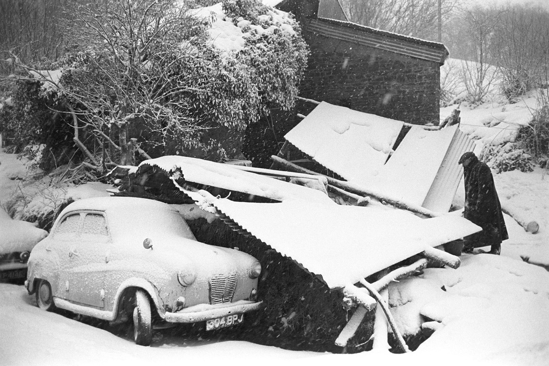 Snow, sheds collapsed under the weight of snow, car. Man looking for ferret under shed. 
[Ivor Brock looking for a ferret under corrugated iron sheds which have collapsed under the weigh of snow at Millhams, Dolton. In the foreground a Baby Austin car is also covered in snow.]