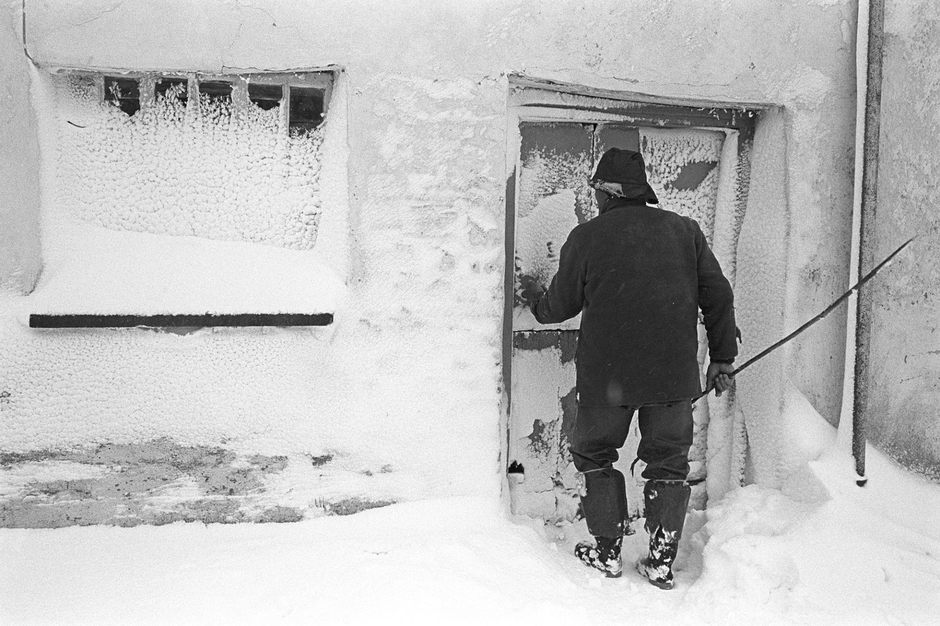Snow, farmer entering farm door after blizzard. 
[Alf Pugsley opening the door of Lower Langham farmhouse in Dolton. He is wearing a sou'wester hat. The door and adjacent window are covered in snow from the blizzard.]