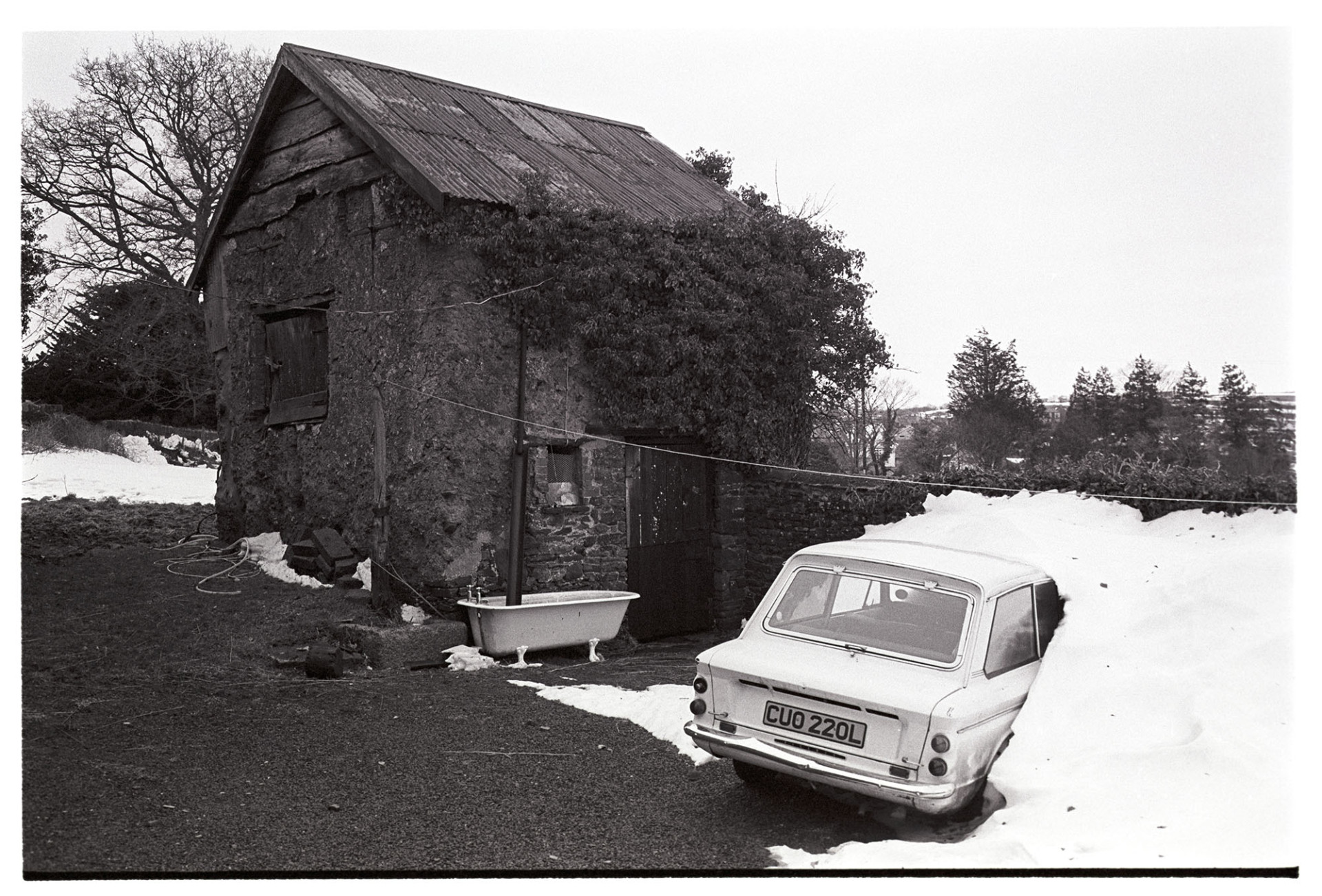 Snow, yard with cob stable barn car in snowdrift, Hillman Imp. 
[A Hillman Imp car covered in a snowdrift at North Ham in Dolton. A stone an corrugated iron barn is in the background, with an old bathtub collecting water from a drainpipe.
See the 'Additional notes by James Ravilious field for more information'.]