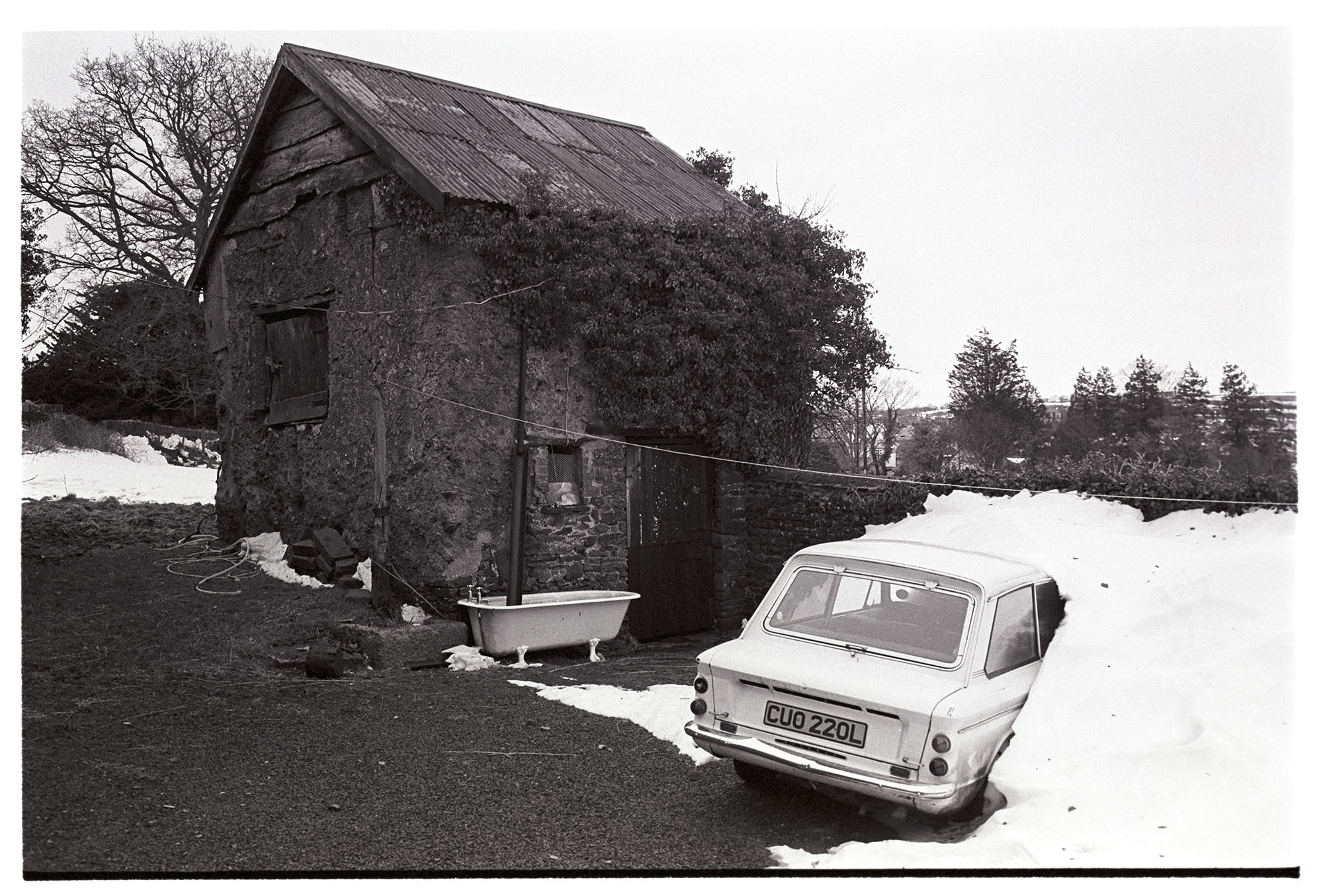 Snow, yard with cob stable barn car in snowdrift, Hillman Imp. <br /> [A Hillman Imp car covered in a snowdrift at North Ham in Dolton. A stone an corrugated iron barn is in the background, with an old bathtub collecting water from a drainpipe.<br /> See the 'Additional notes by James Ravilious field for more information'.]