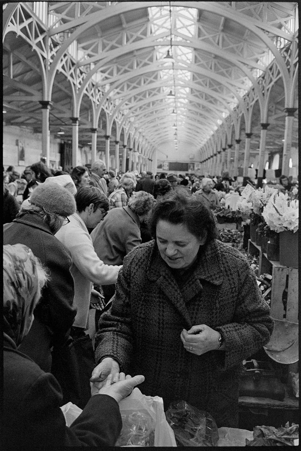 Stalls at Pannier market, eggs, vegetables, flowers. 
[A woman buying goods from a stall at Barnstpale Pannier Market. Other stallholders and shoppers can be seen in the background, as well as buckets of flowers for sale on one of the stalls.]