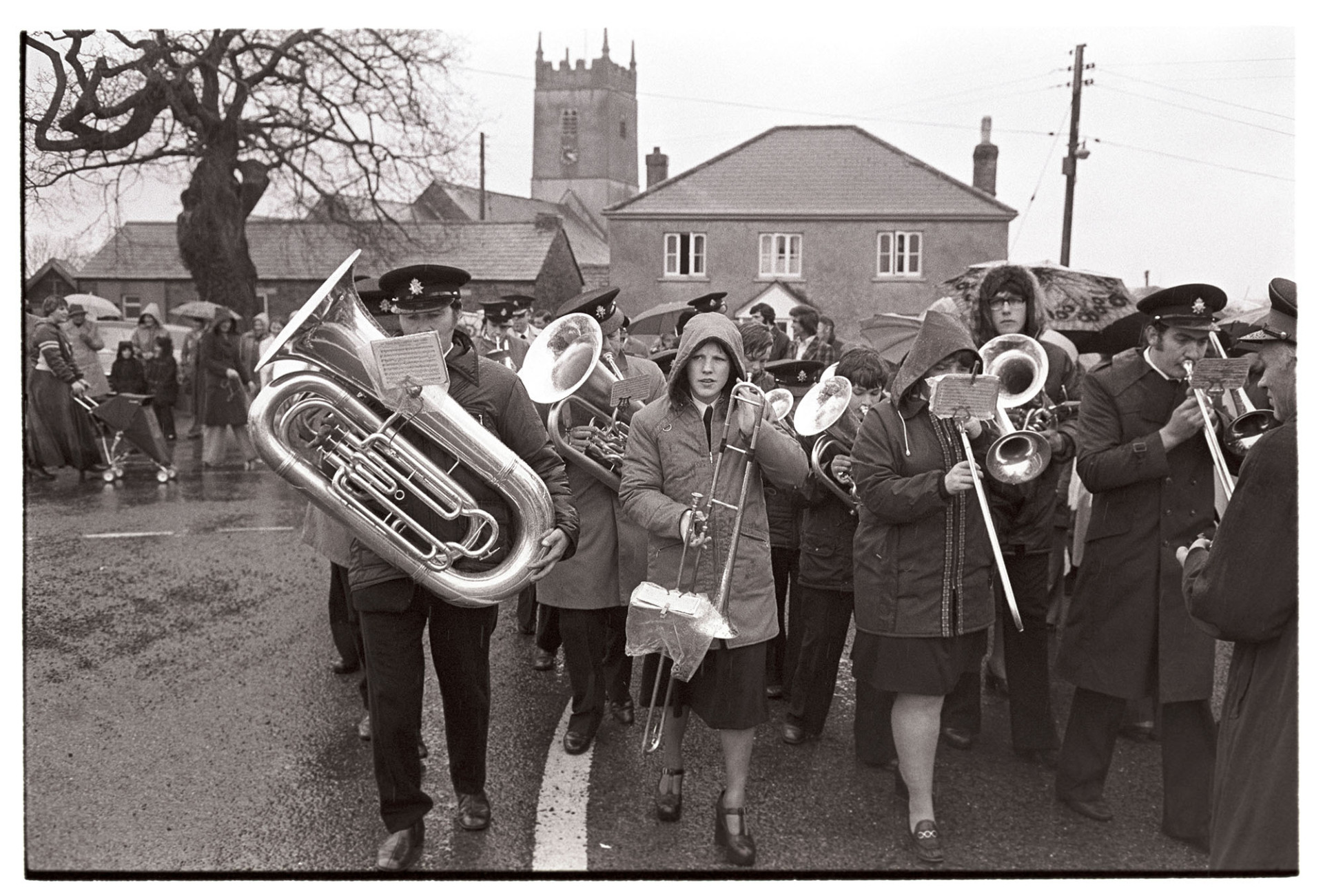 Brass band at carnival parade. 
[A brass band performing in the rain at Shebbear Carnival. The band might be Northlew Brass Band. Houses and the church tower is visible in the background.]
