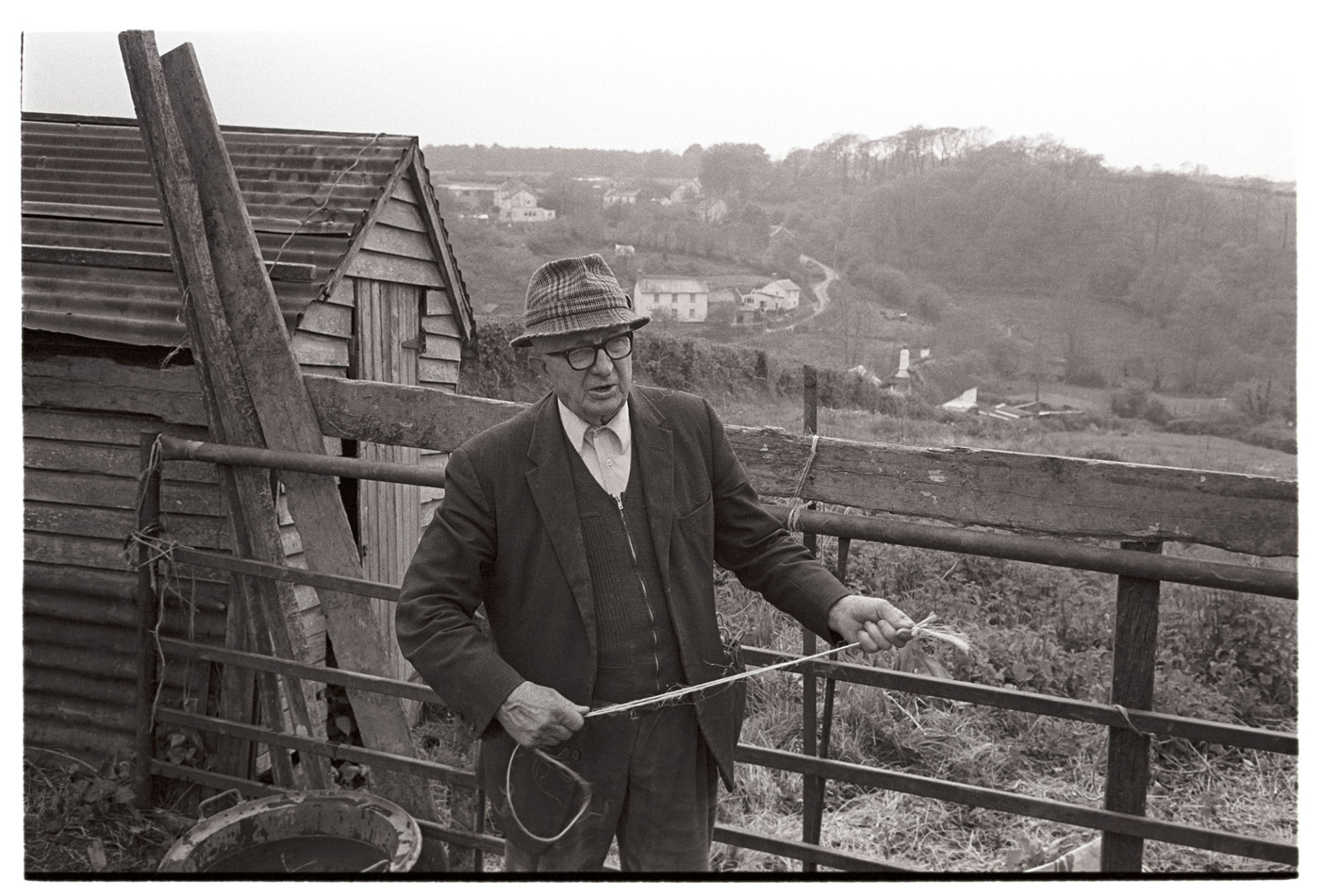 Farmer chatting to me [James Ravilious].
[Mr Parkhouse stood in front of a field gate and wooden barn with a corrugated iron roof at Hollocombe. The village can be seen in the distance. He is also holding a piece of twine.]