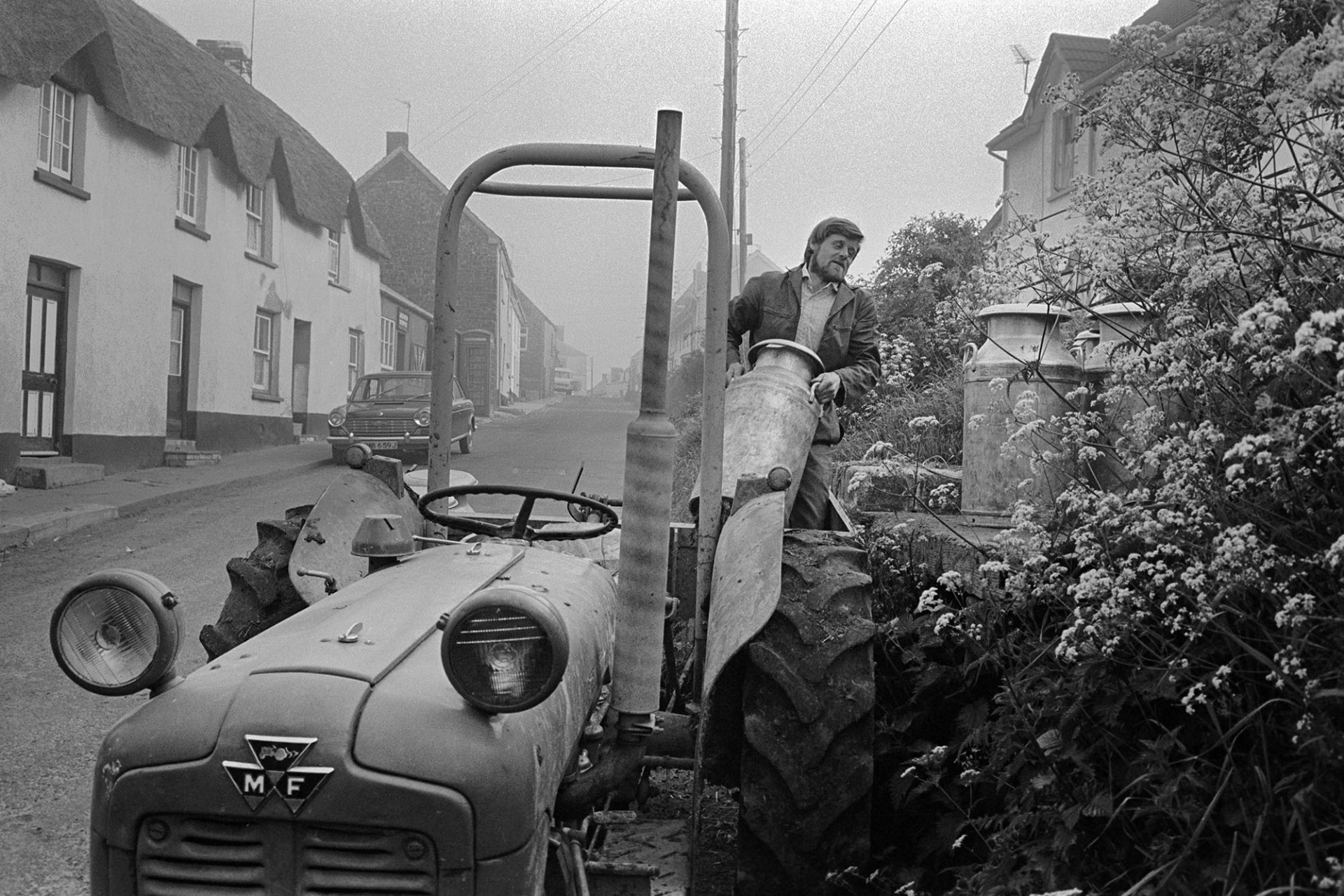 Farmer transferring milk churns to stand in village. 
[A man lifting milk churns from his tractor and link box onto a milk churn stand in the village of Sheepwash. Thatched cottages and a parked car are visible in the street.]