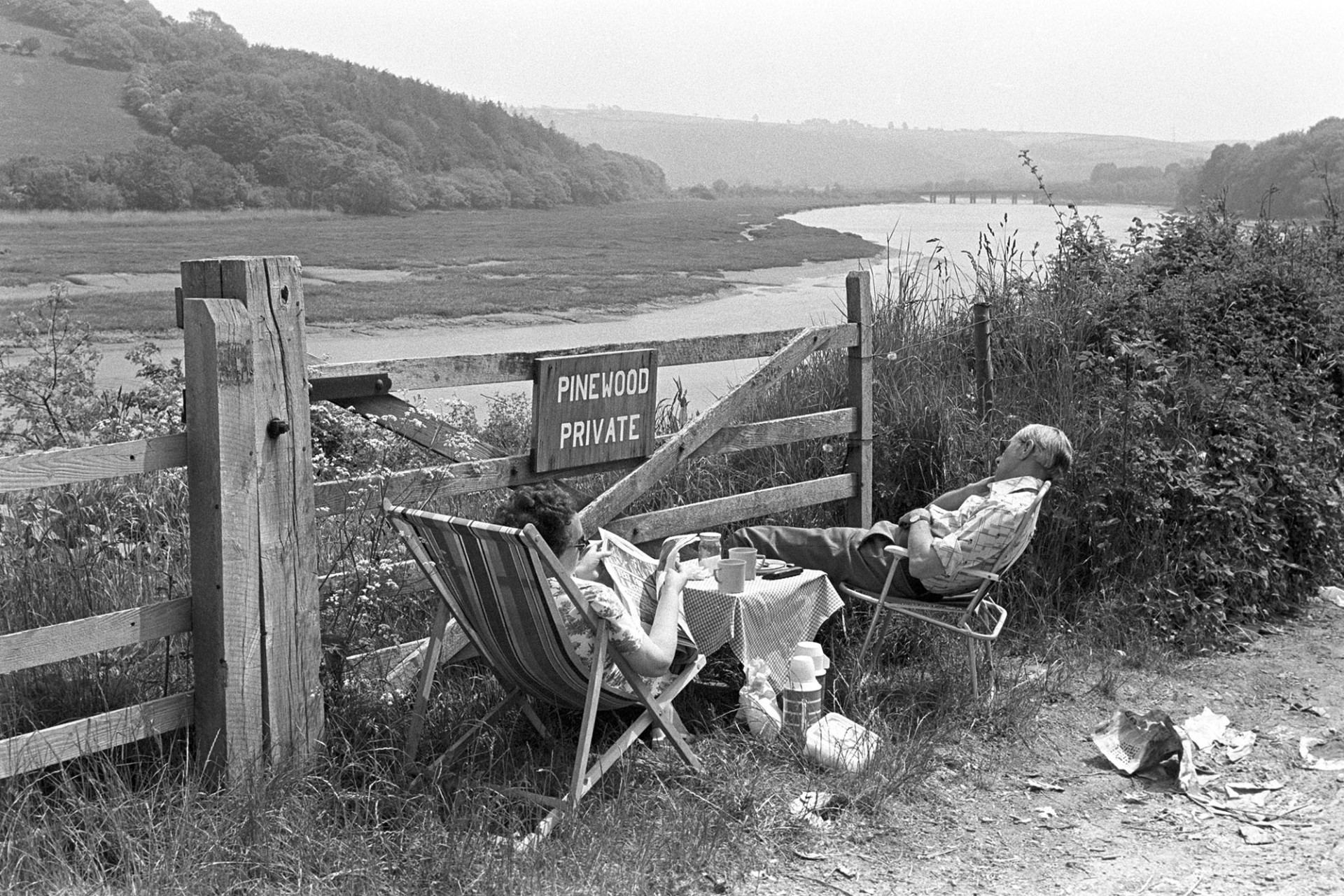Sleeping tourists after picnic beside estuary. 
[Two tourists sat on picnic chairs behind a wooden gate by the river Torridge estuary at Bideford. One is sleeping and the other is reading a newspaper. They also have a small picnic table and thermos flasks.]