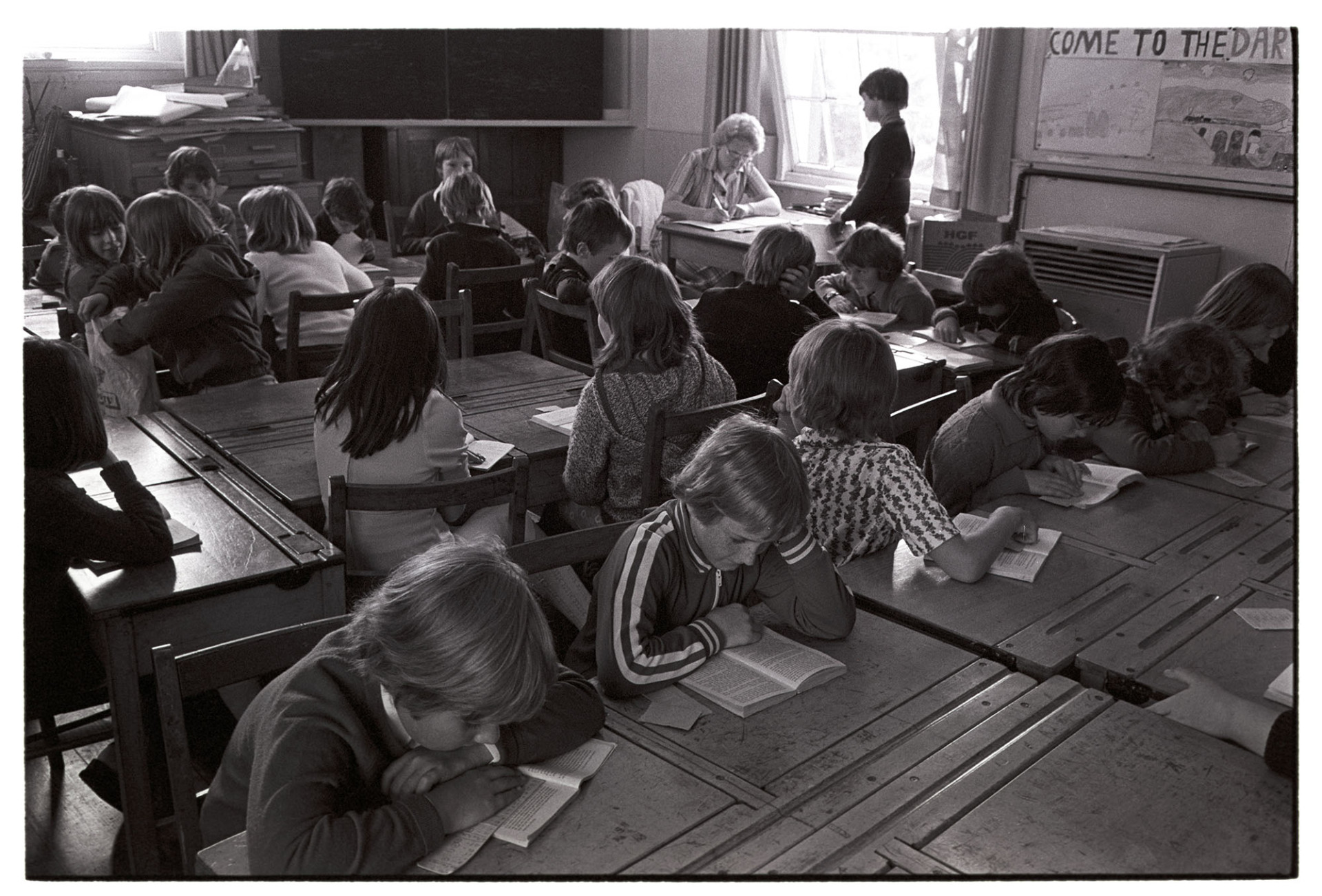 Children in class working reading.
[Children in a classroom sitting at desks and reading. A teacher is sitting by the window checking a child's work, at Blue Coats School, Torrington.]