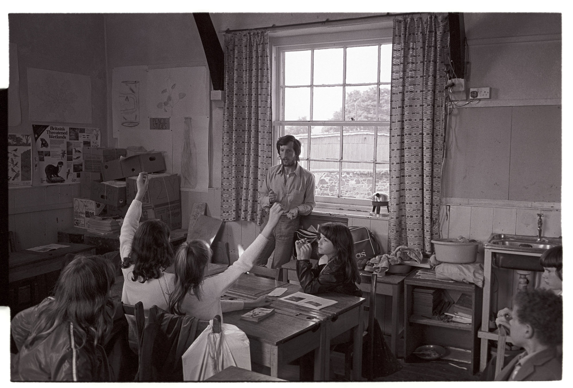 Schoolchildren and teacher in class, answering questions.
[Children in a classroom sitting at desks and holding up hands to answer questions.  A teacher is standing by a window in the classroom and talking, at Blue Coats School Torrington.]