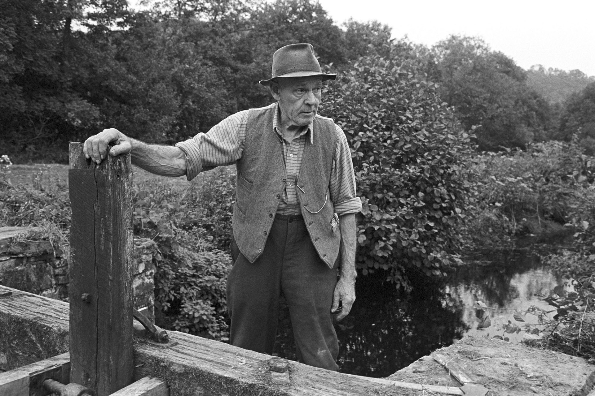 Man, repair of sluice gate of mill leat. 
[Herbert Snell stood by a wooden sluice gate he has been repairing on a leat near Head Mill, Kings Nympton.]