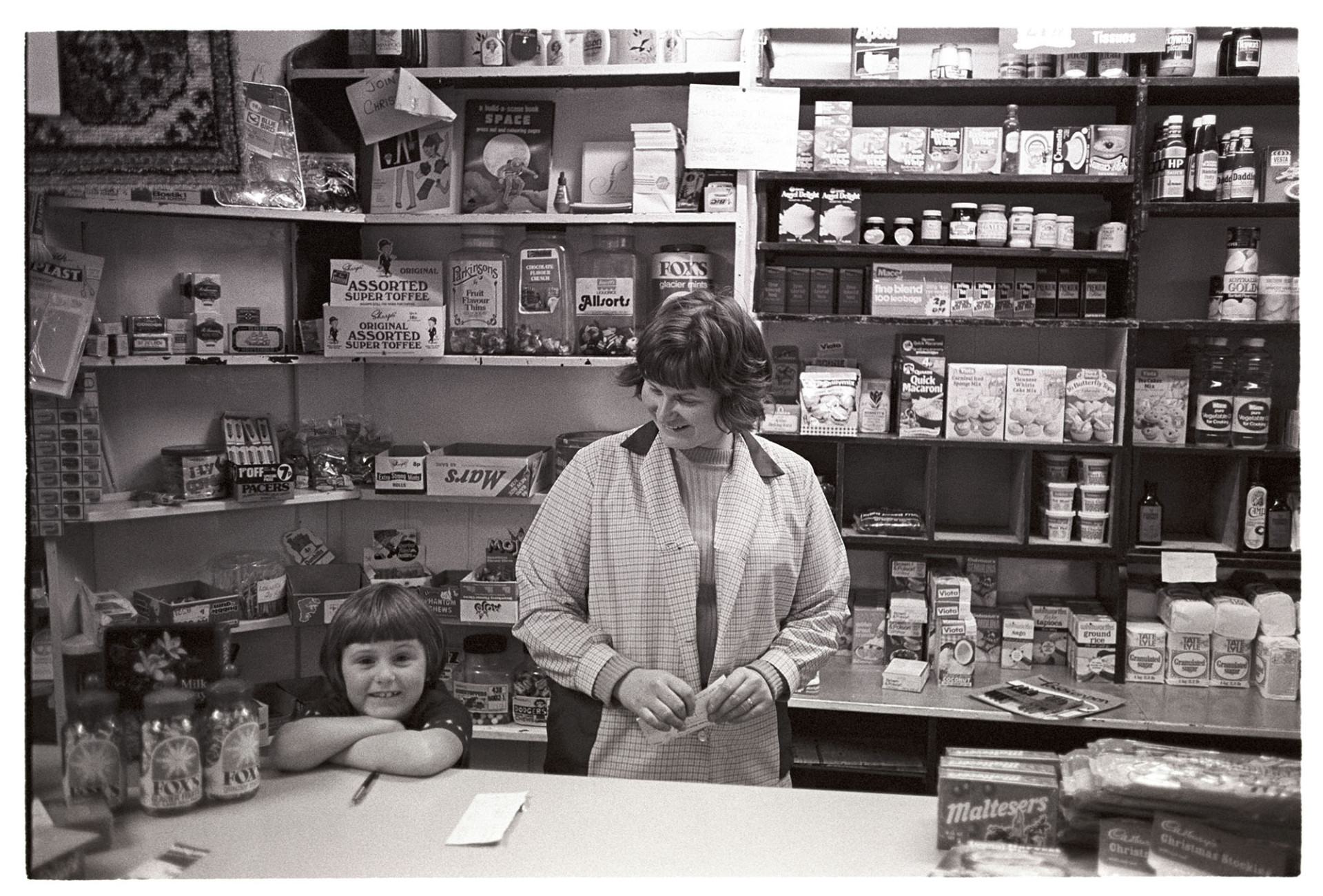 Interior of village shop with woman proprietor and daughter. 
[Christine Keogh, the proprietor of the General Stores at Merton, and her daughter stood behind the counter of the shop. Various good are displayed on shelves behind the counter, including sweets, teabags and cupcake mixes.]